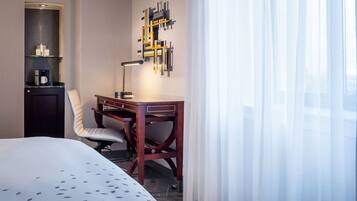 A hotel room with a bed, a desk with a lamp, a chair, a shelf with a coffee maker, and a wall art piece.