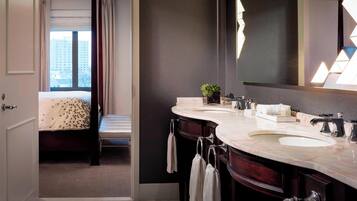 A modern hotel bathroom with two sinks, a large mirror, and a bed with a view of a cityscape.