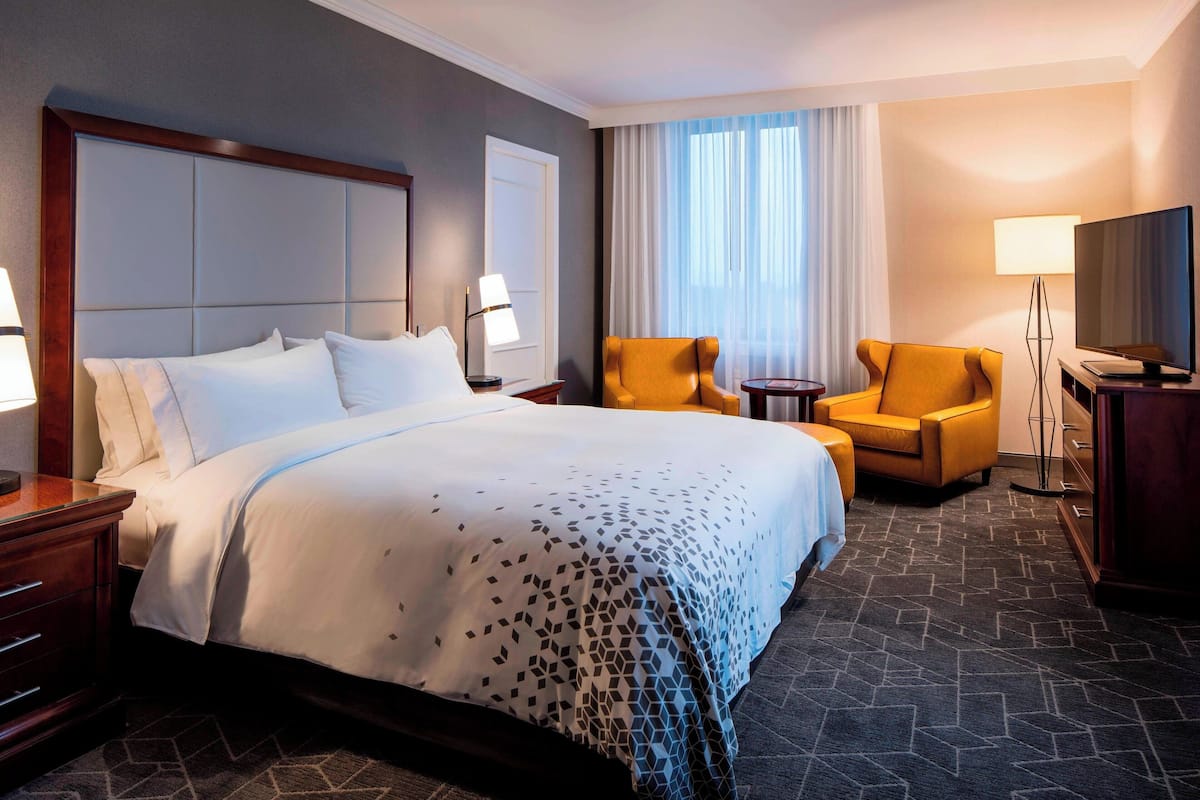 A hotel room with a large bed, two chairs, a desk with a TV, and a window with curtains.