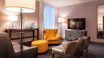 Ein Hotelzimmer mit Sofa, Sessel, Hocker, Fernseher und Schreibtisch mit Lampe.