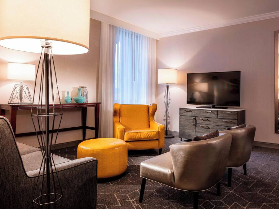 Ein Hotelzimmer mit Sofa, Sessel, Hocker, Fernseher und Schreibtisch mit Lampe.