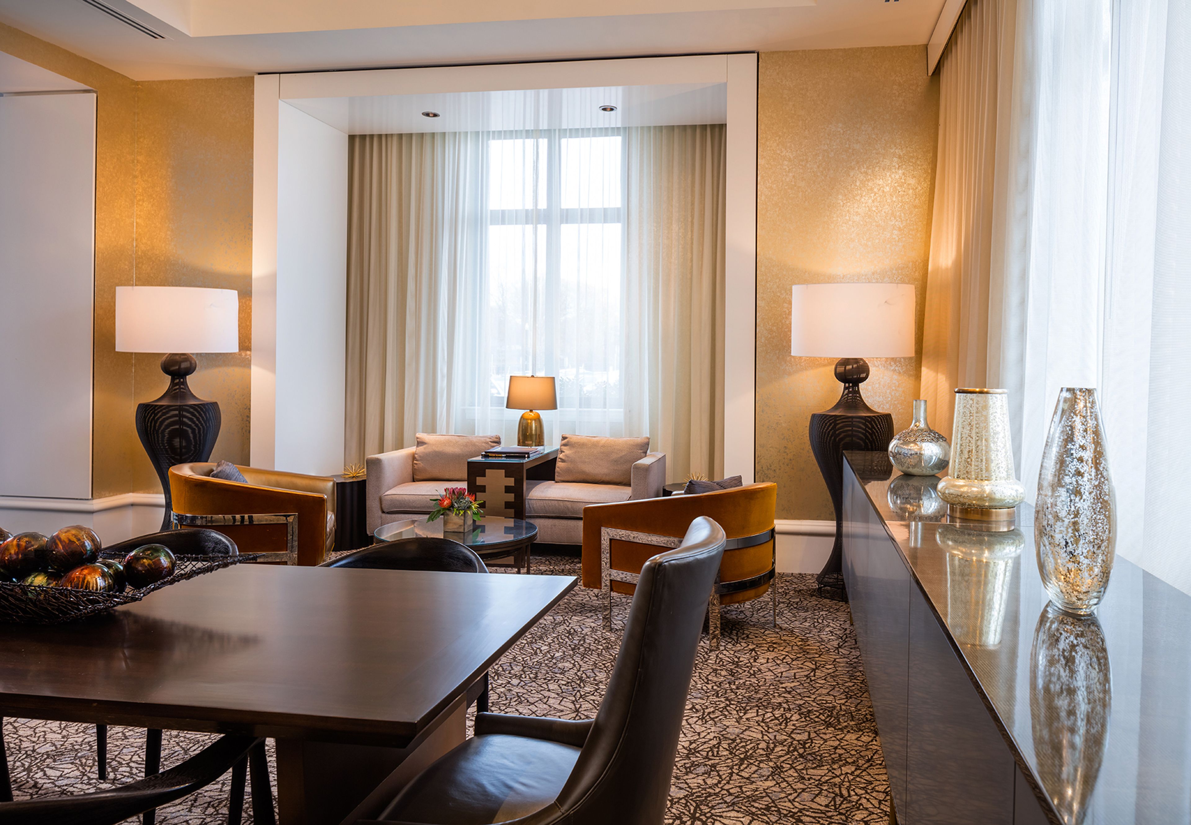 A hotel room with a dining area, a sofa set, two lamps, and a large window with curtains.
