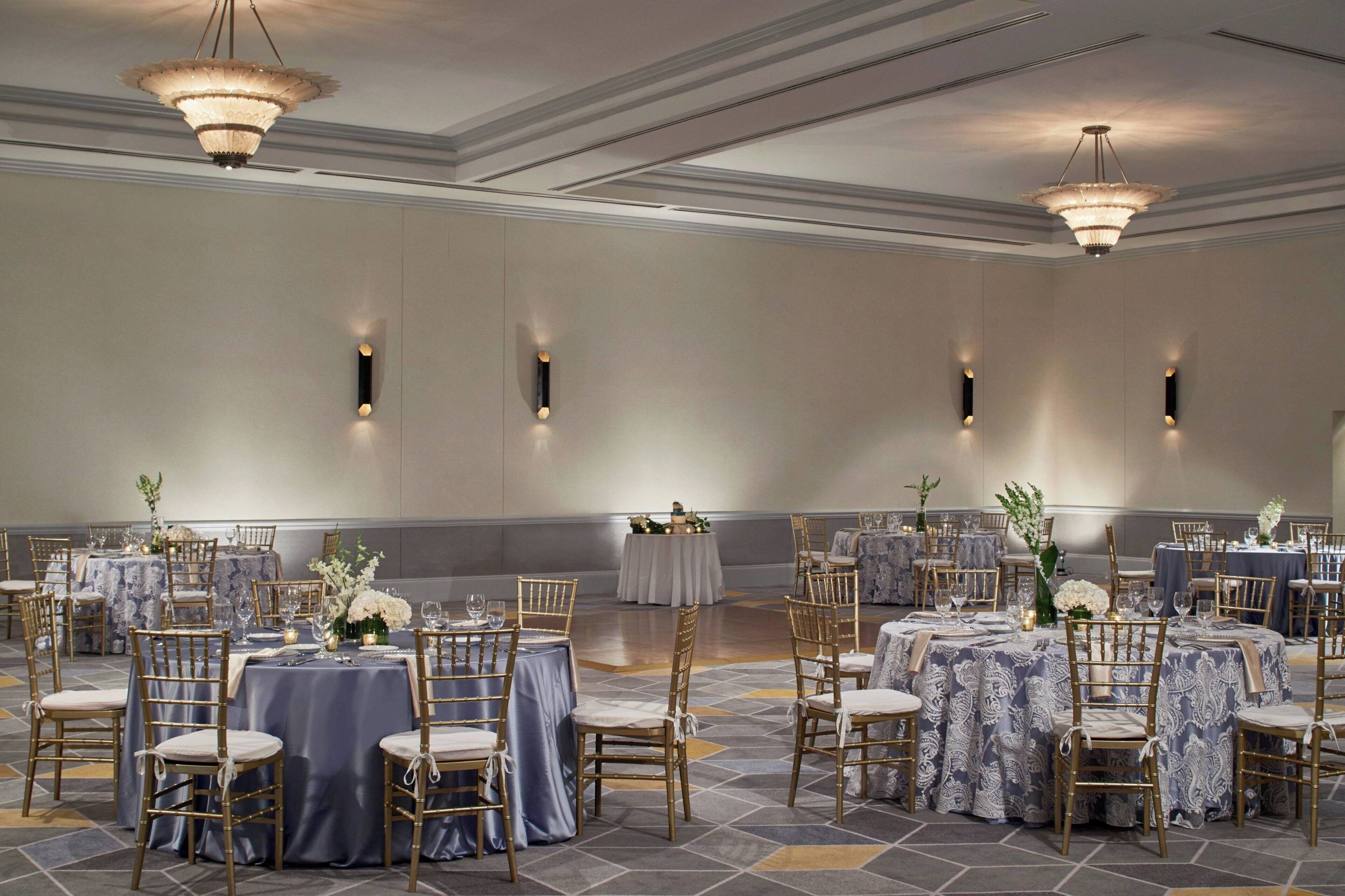 A banquet hall with round tables, gold chairs, and chandeliers.