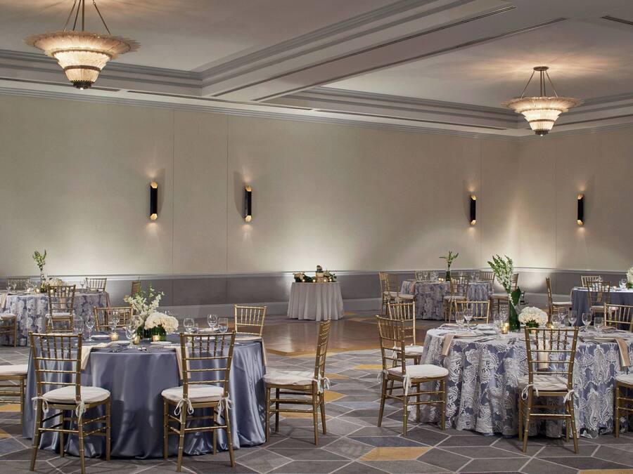 A banquet hall with round tables, gold chairs, and chandeliers.