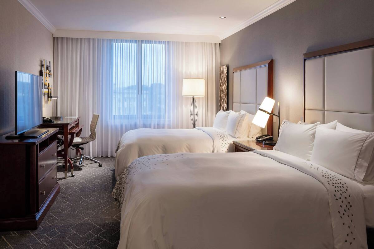 A hotel room with two beds, a desk with a computer, a chair, a lamp, and a large window with curtains.