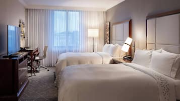 Habitación de hotel con dos camas, un escritorio con computadora, una silla, una lámpara y un ventanal grande con cortinas.