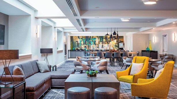 Um hall de hotel moderno com um sofá, poltronas, uma mesa de centro e uma área de refeições com mesas e cadeiras.