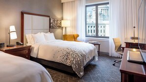 Quarto de hotel com uma cama grande, uma escrivaninha, uma cadeira, um abajur e vista para um edifício pela janela.