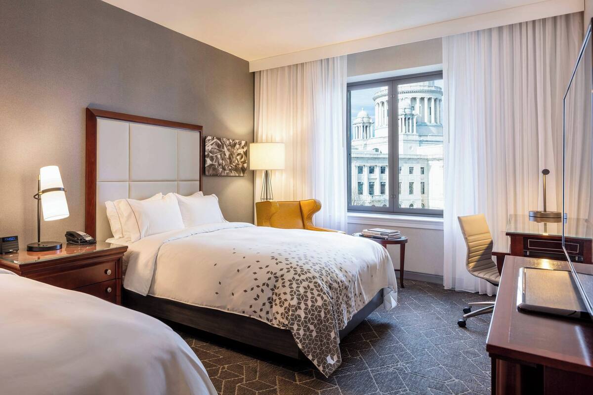 A hotel room with a large bed, a desk, a chair, a lamp, and a view of a building through the window.