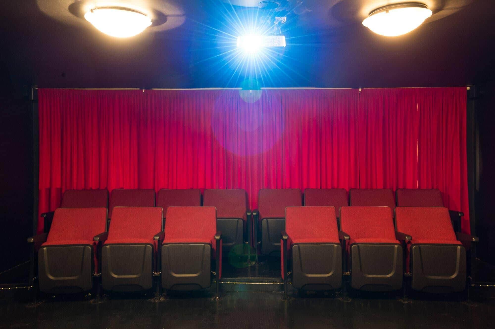 Un teatro con cortinas rojas, hileras de asientos negros y dos luces en el techo.
