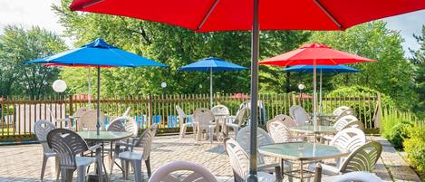 An outdoor patio with tables and chairs under red and blue umbrellas, surrounded by a wooden fence and greenery.