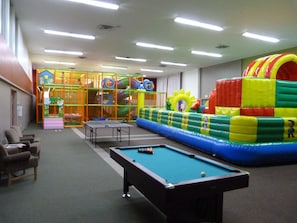 Un espacio recreativo interior con mesa de billar, asientos y zona de juegos con estructuras inflables.