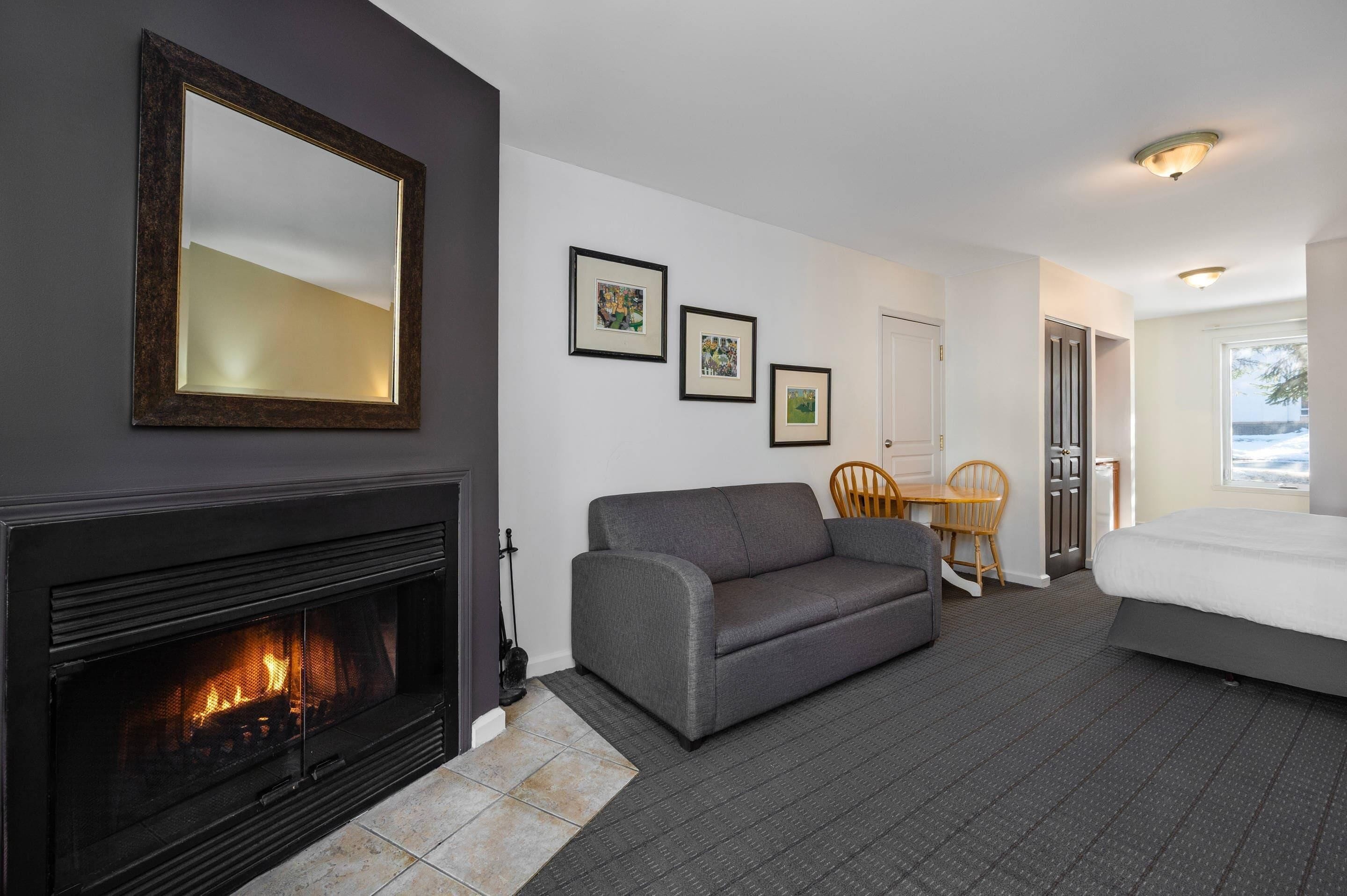 A hotel room with a fireplace, a sofa, a bed, a dining table with chairs, and framed pictures on the wall.