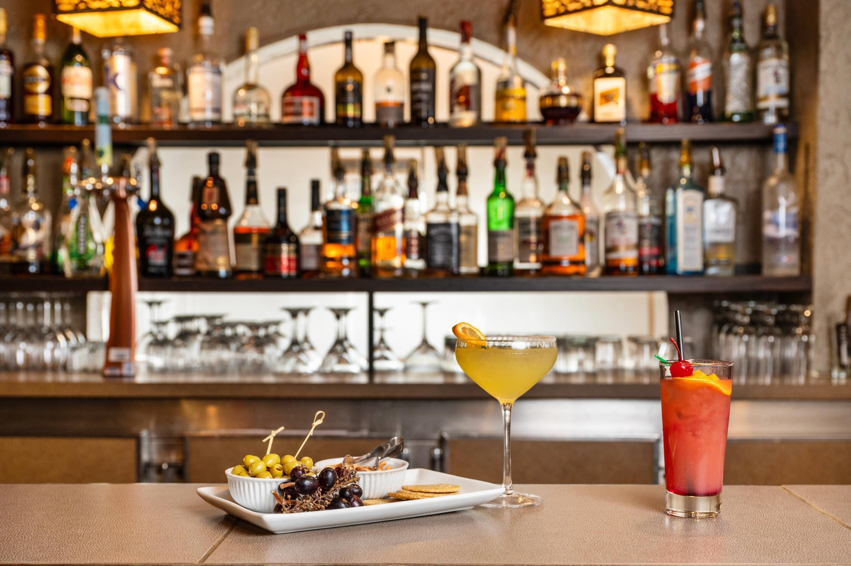 Zona de bar con diversas botellas de licor, una copa de martini, una copa de cóctel y un plato de aceitunas y galletas.