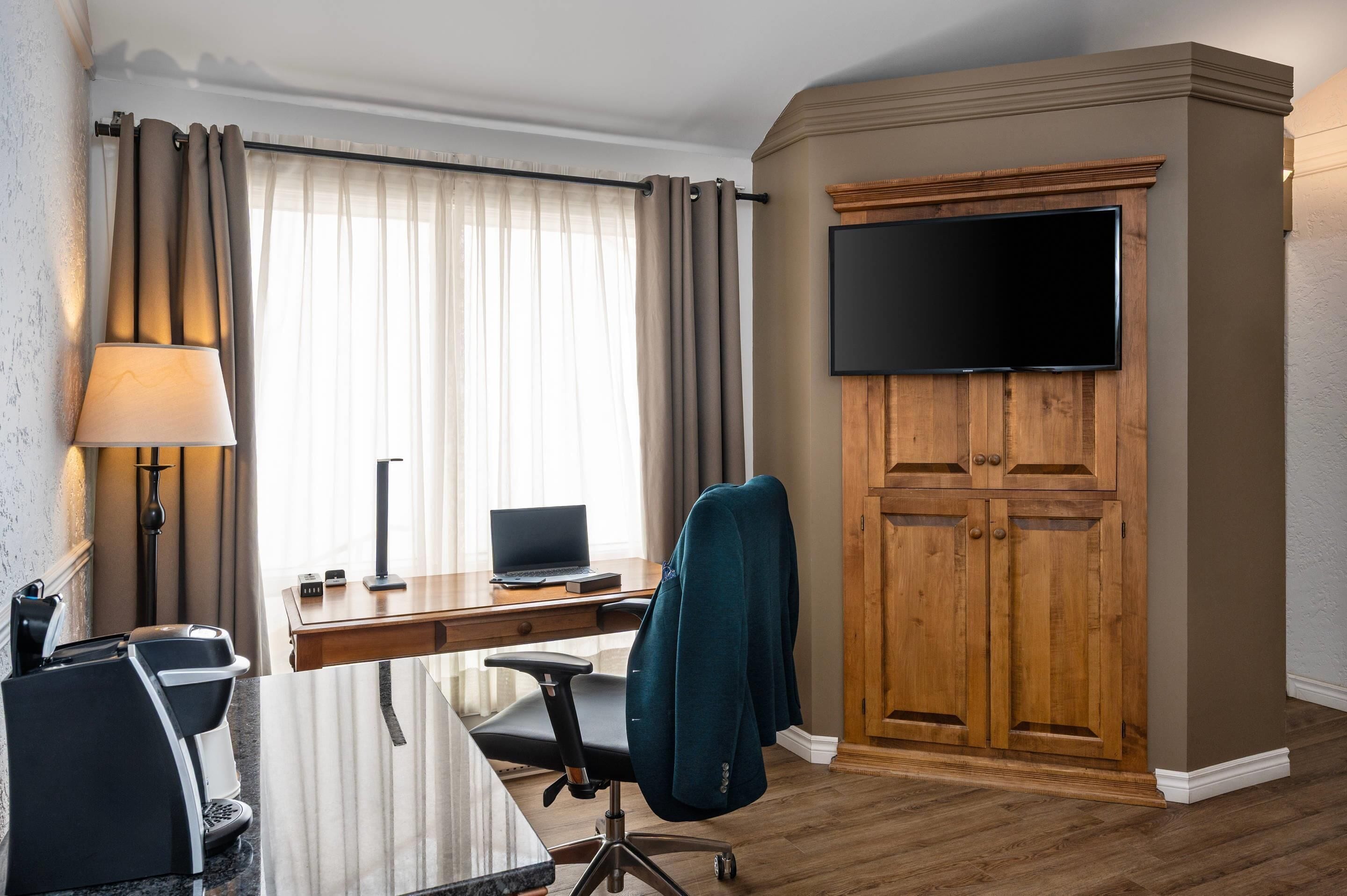 Une chambre d’hôtel avec un bureau en bois, une chaise, un téléviseur fixé à un meuble en bois, une lampe et une fenêtre avec des rideaux.