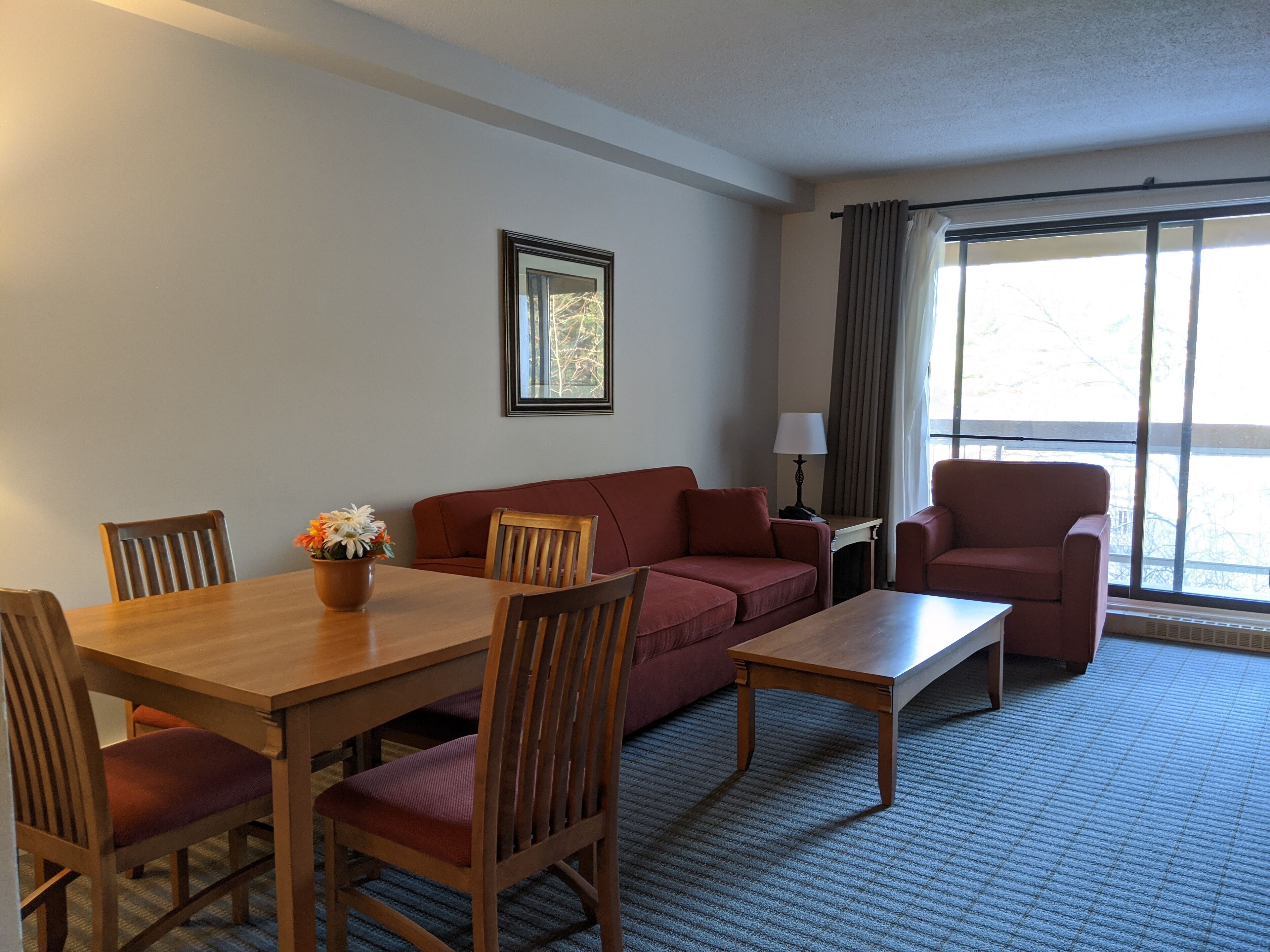 A hotel room with a wooden dining table, red sofa, and two armchairs. There is a framed picture on the wall and a window with curtains.