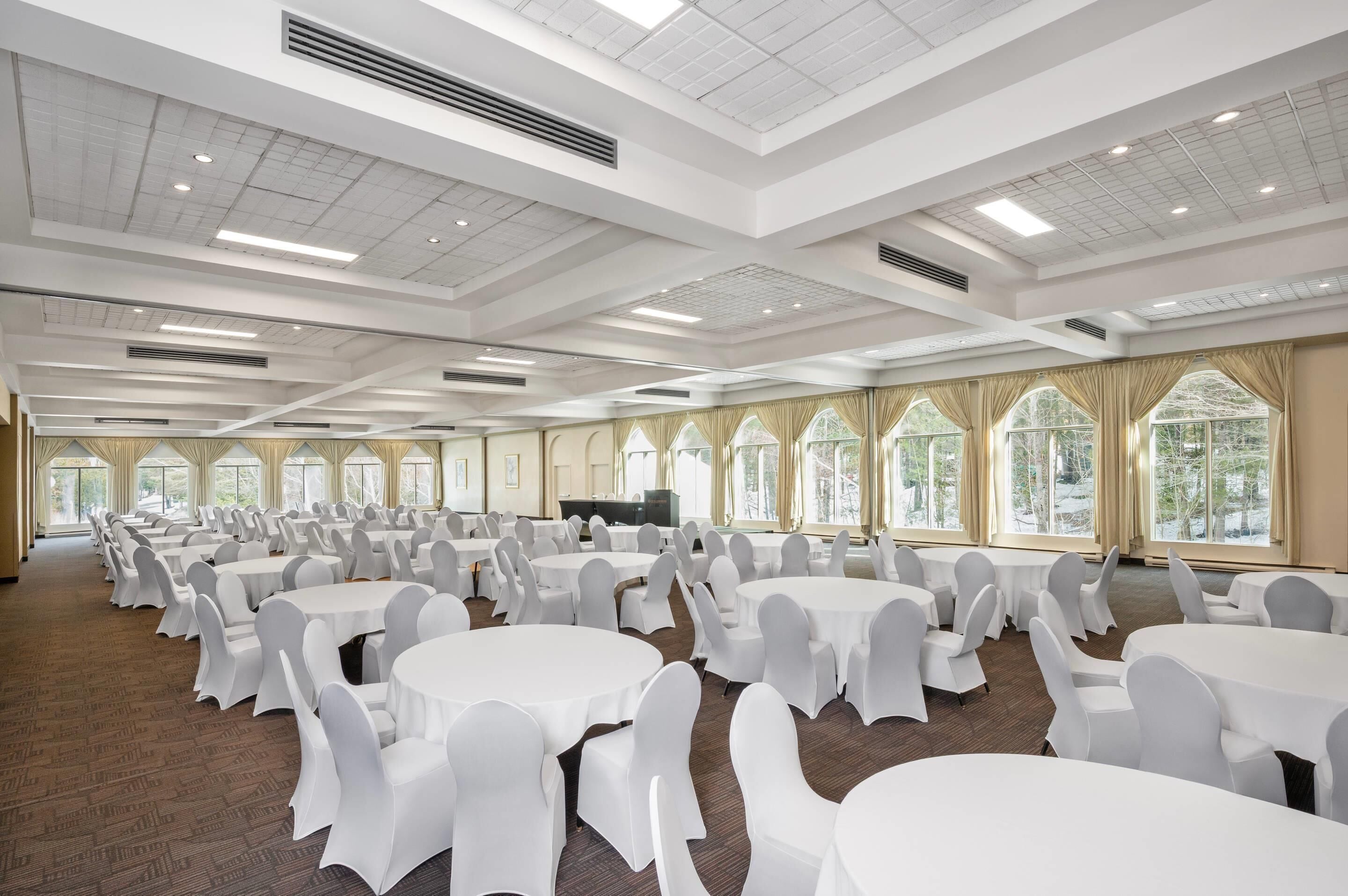 A large conference room with multiple round tables and chairs, a high ceiling with grid-like patterns, and large windows with curtains.