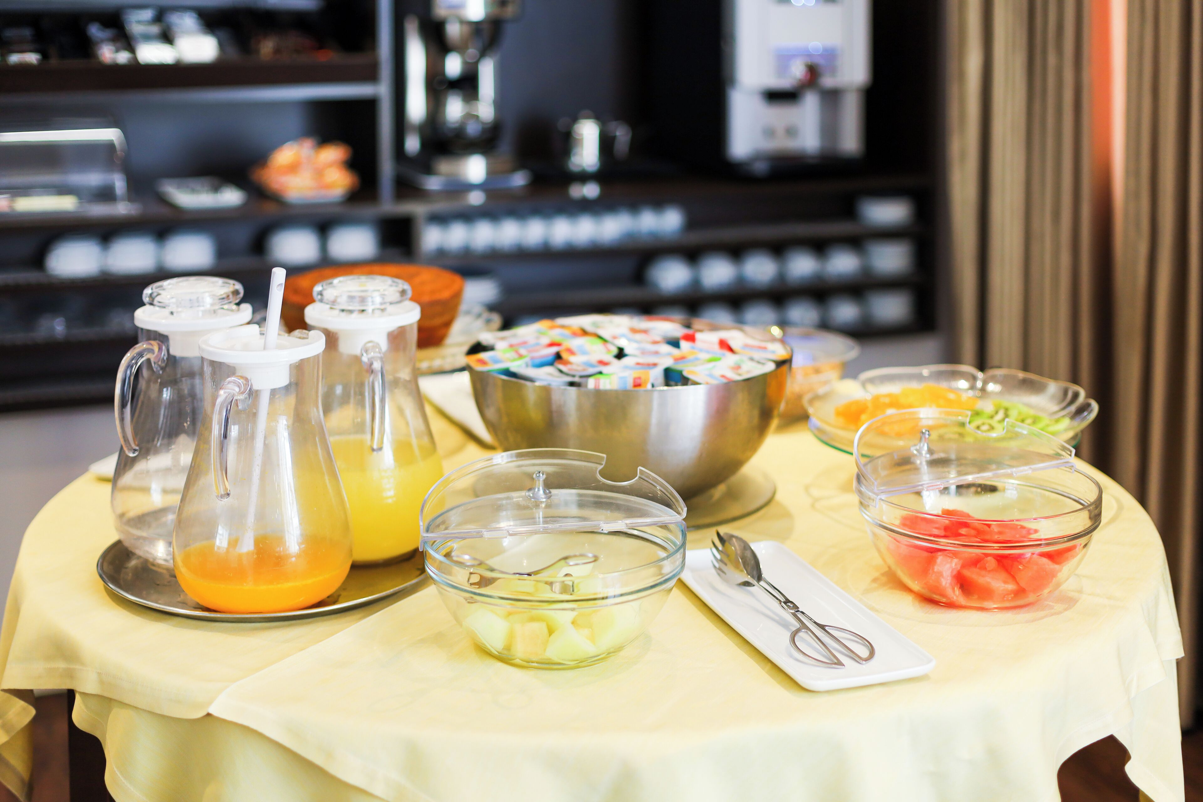 A buffet table with various food items and beverages, including a bowl of fruit, a pitcher of orange juice, and a bowl of cereal.