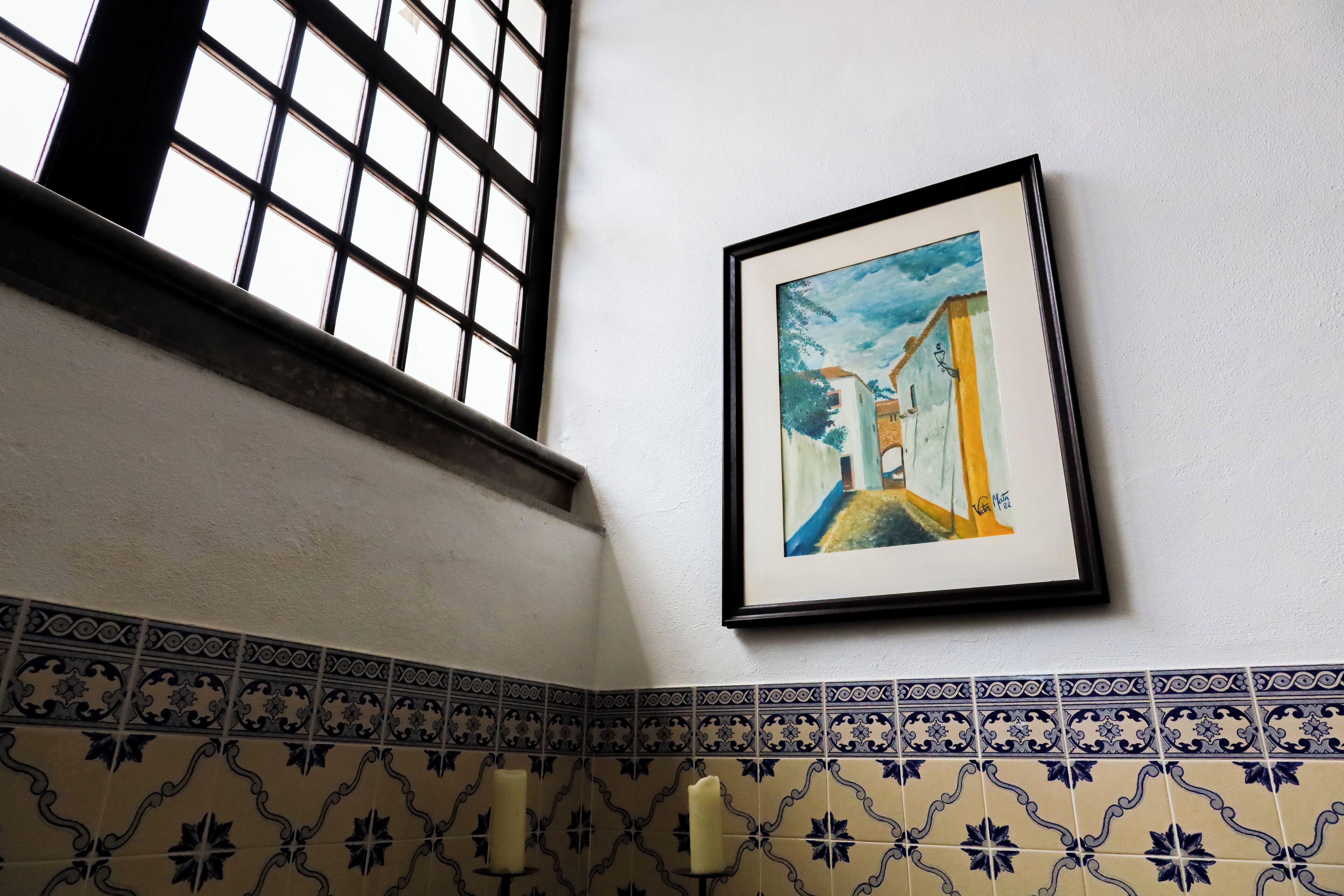 A framed painting of a street scene on a wall, a window with a grid pattern, and patterned tile flooring.