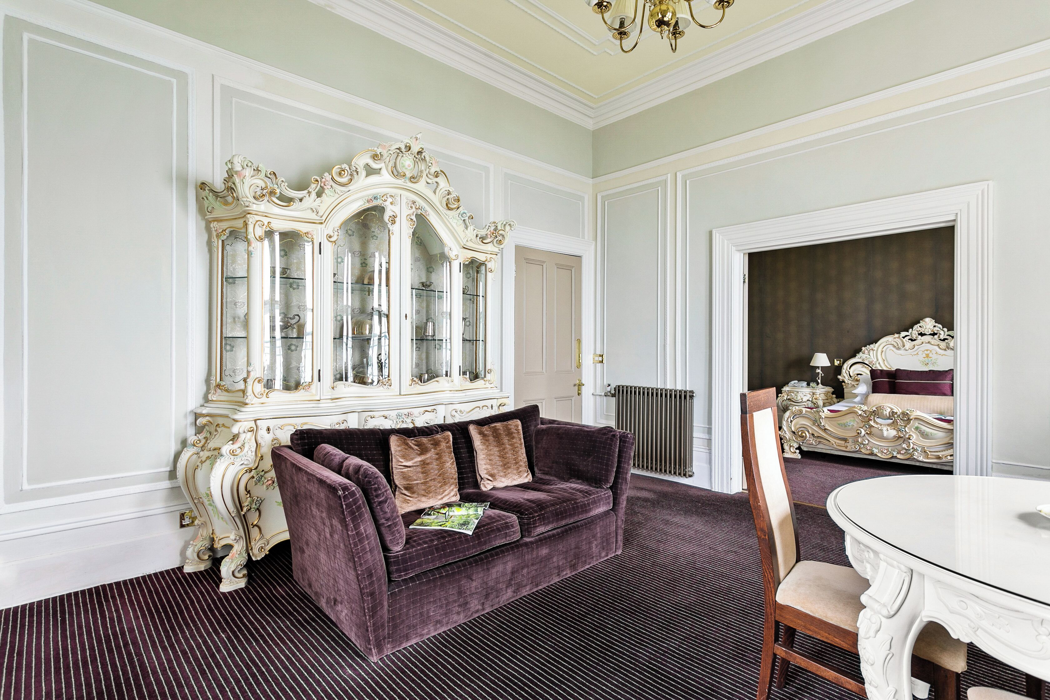 A room with a purple velvet sofa, a white ornate cabinet, and a round dining table.