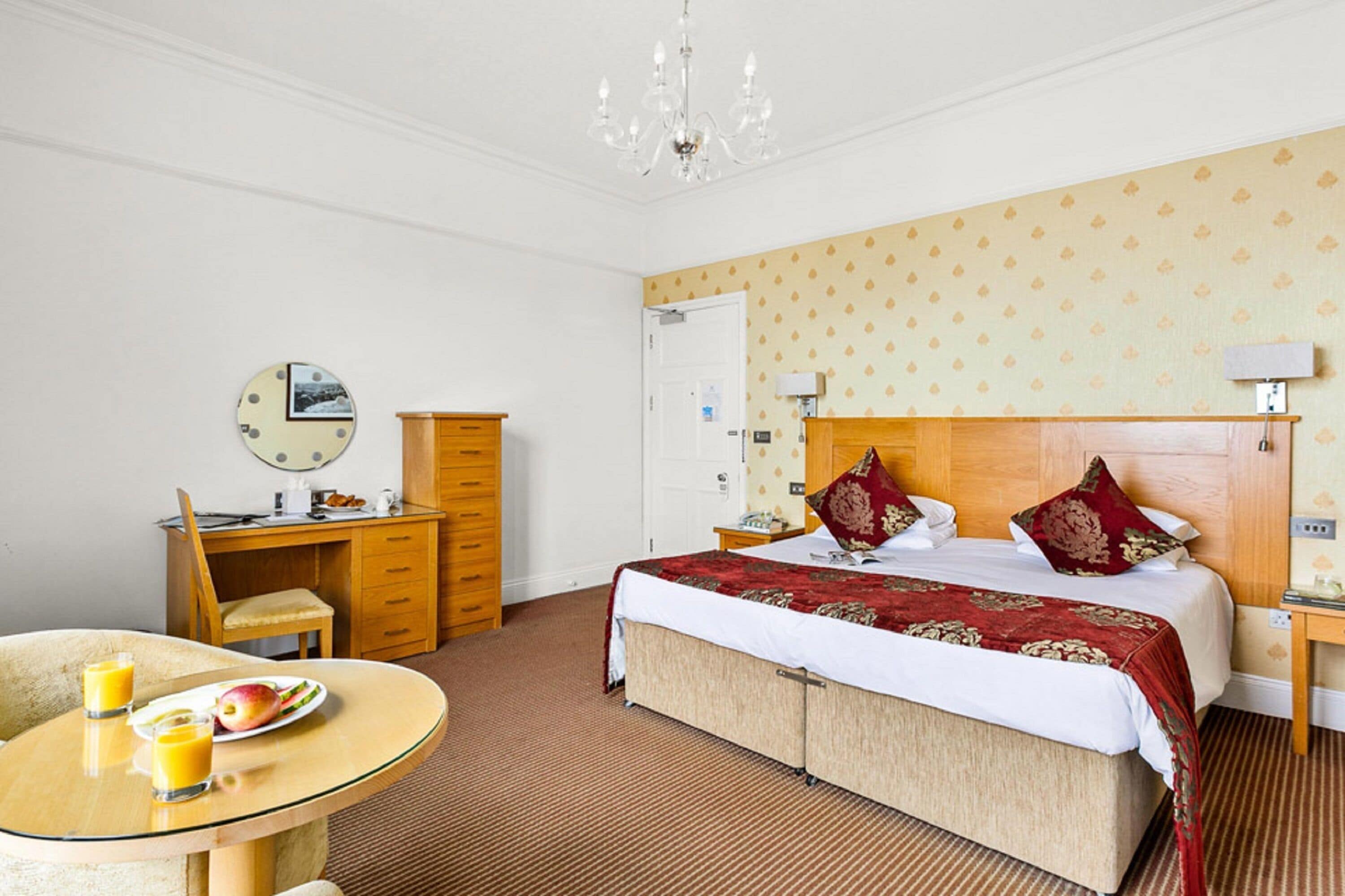 A hotel room with a bed, a desk, a chair, a table with breakfast, and a chandelier.