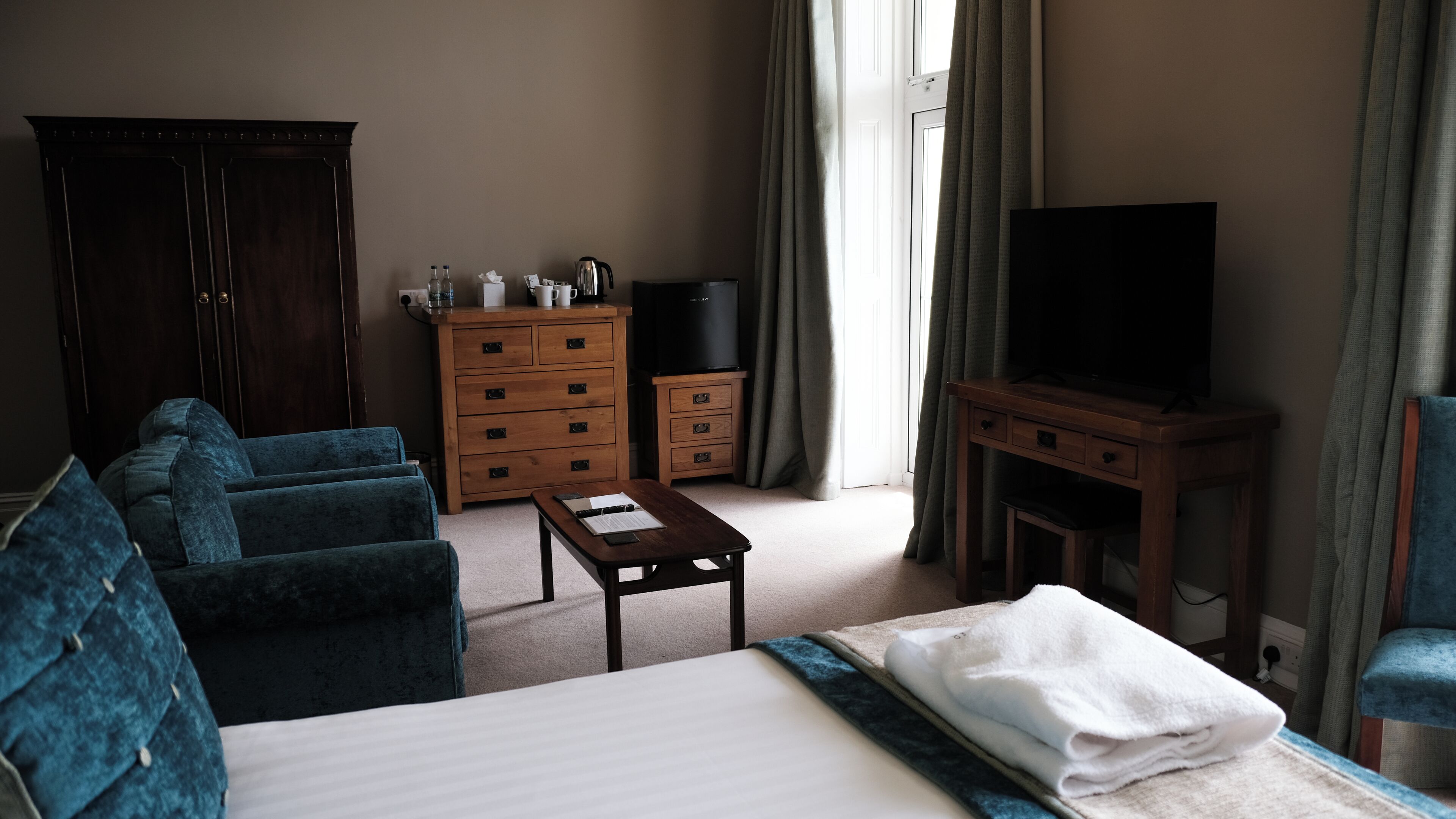 A hotel room with a bed, a blue sofa, a wooden dresser, a television, and a window with curtains.