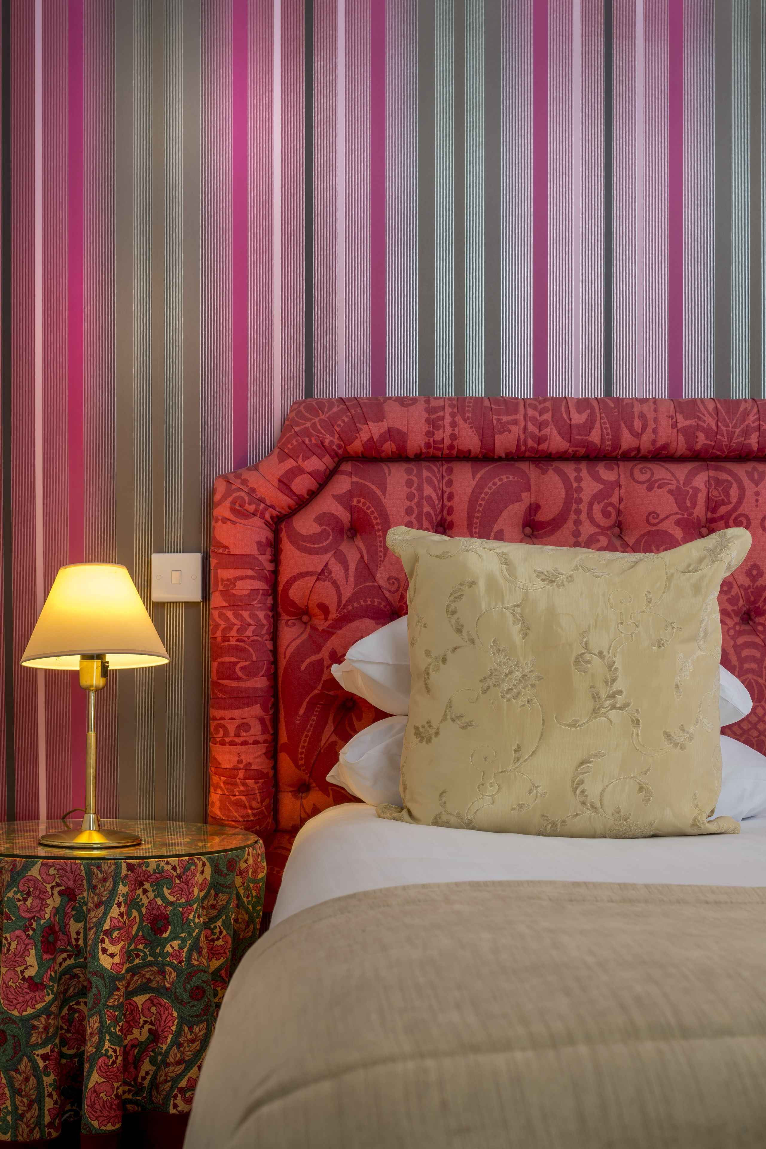 Een hotelkamer met een gestreepte muur, een gewatteerd hoofdeinde, een nachtkastje met dessin en een lamp.