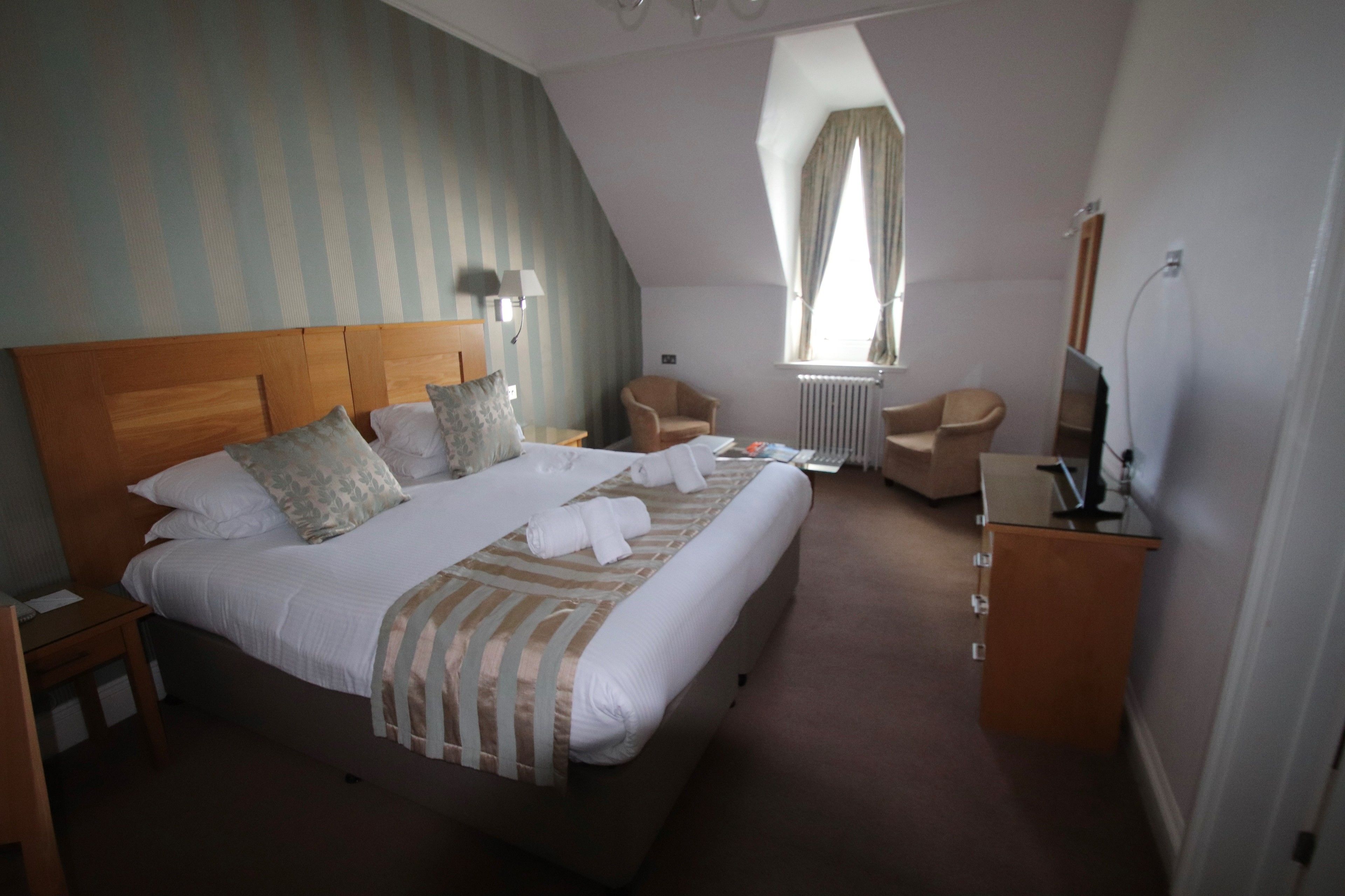 Een hotelkamer met een groot bed, twee fauteuils, een bureau met een computer en een raam met gordijnen.