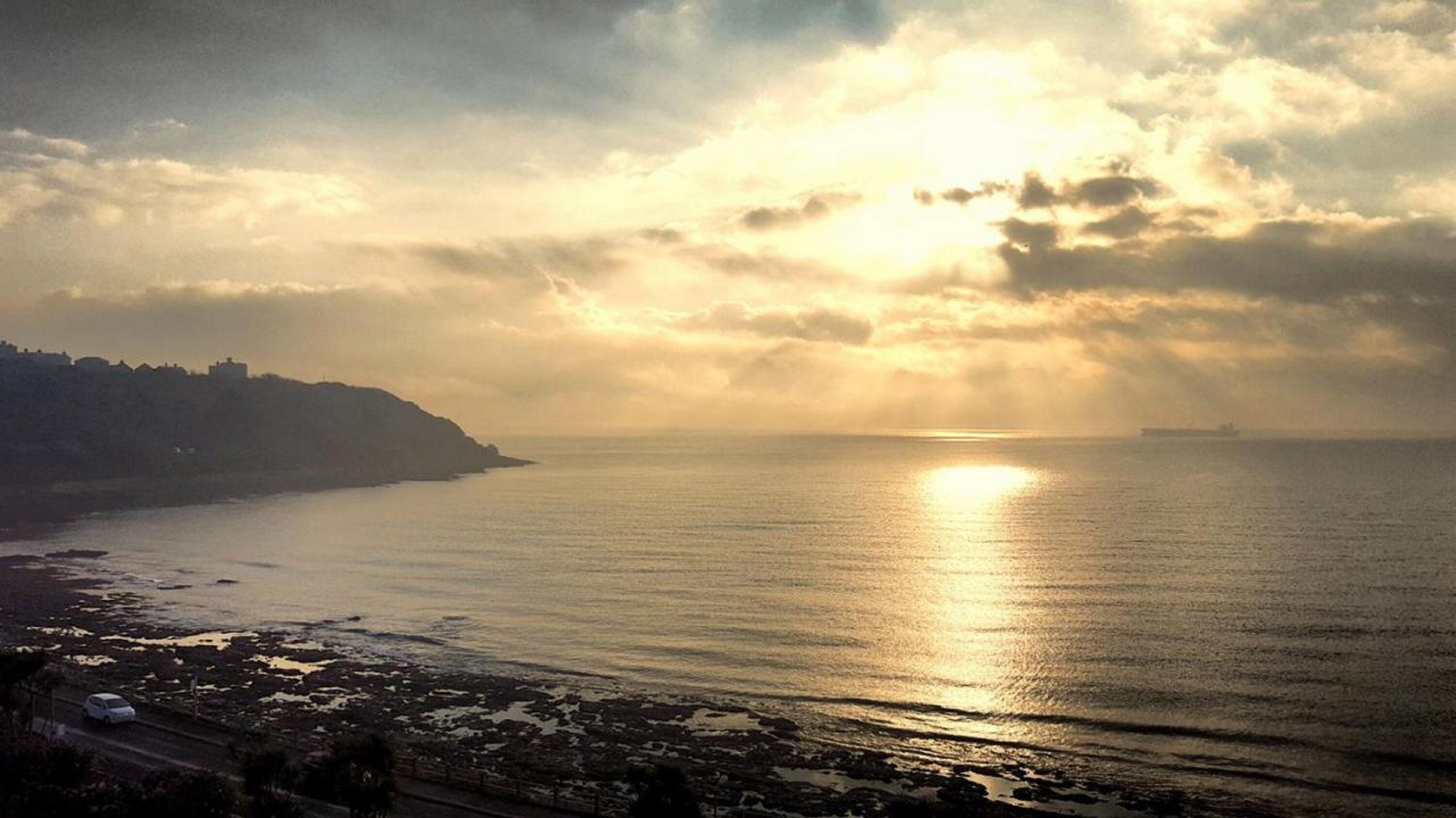A coastal scene with a road leading to a beach, a body of water reflecting sunlight, and a distant city skyline.