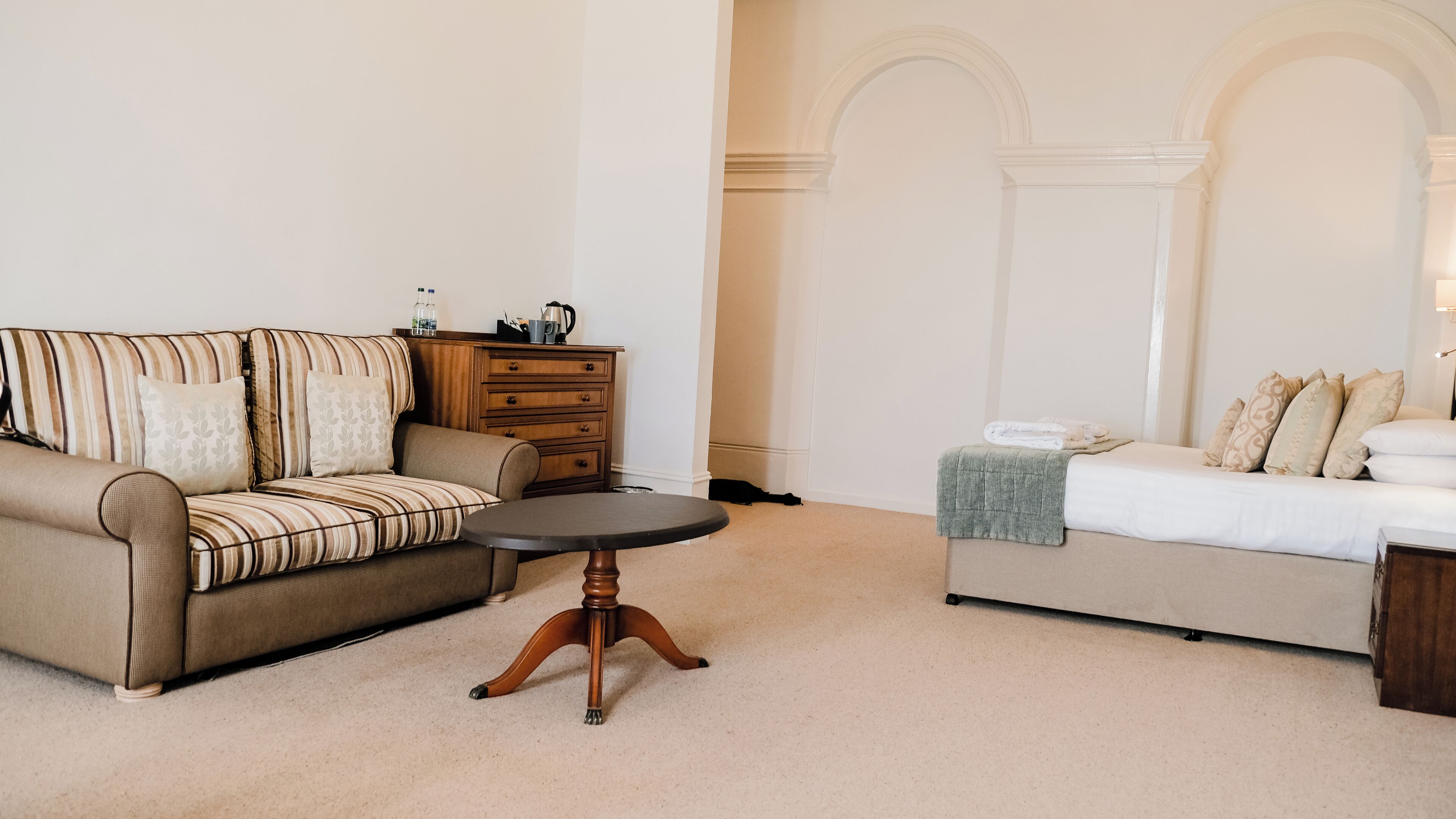 Un salon avec un canapé à rayures, un meuble en bois, une table ronde et un lit doté d’un matelas blanc et d’oreillers beiges.