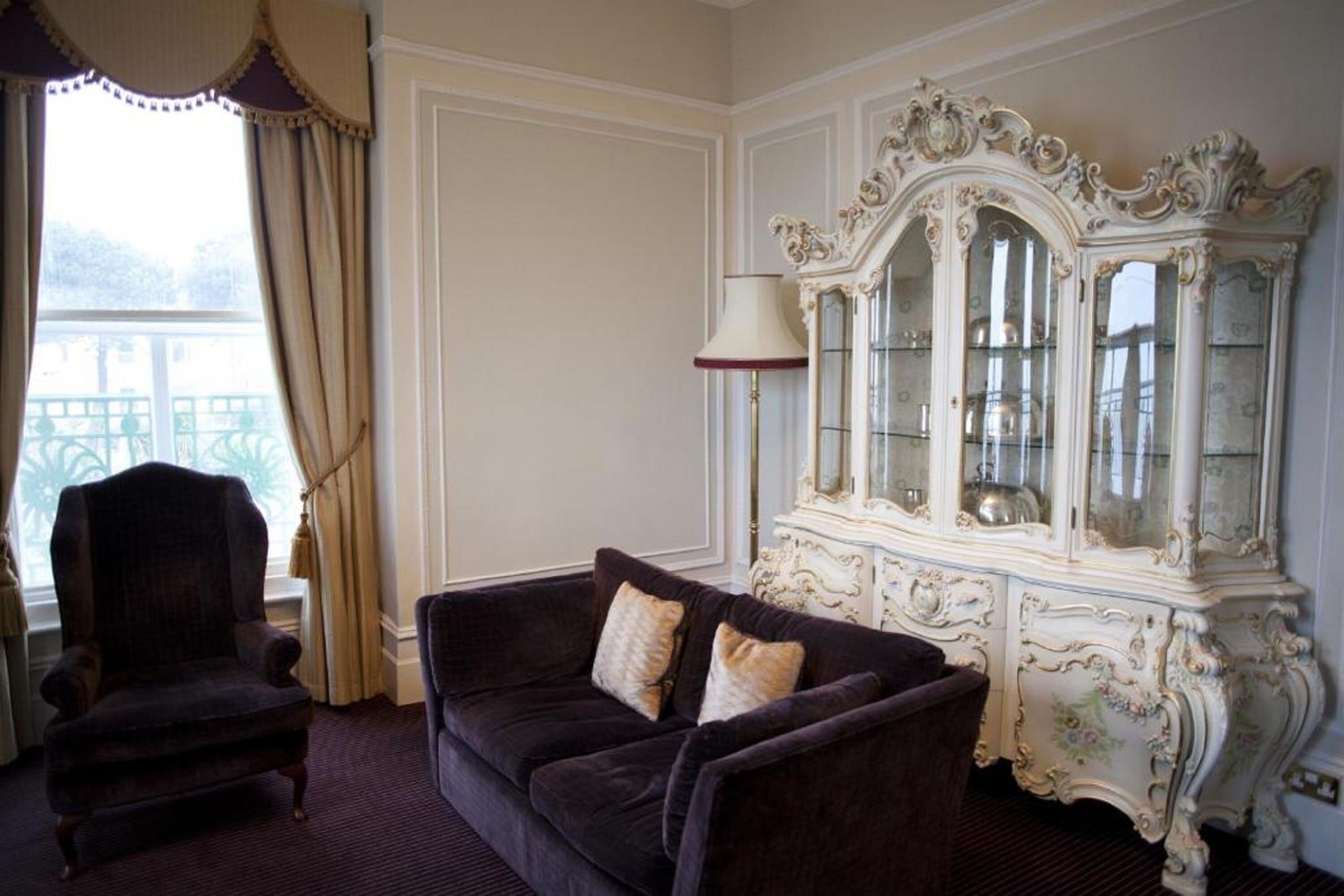 A room with a dark velvet sofa, a white ornate cabinet, and a window with curtains.