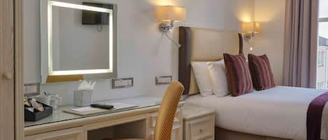 A hotel room with a bed, bedside lamps, a desk with a chair, a mirror, and a window with curtains.