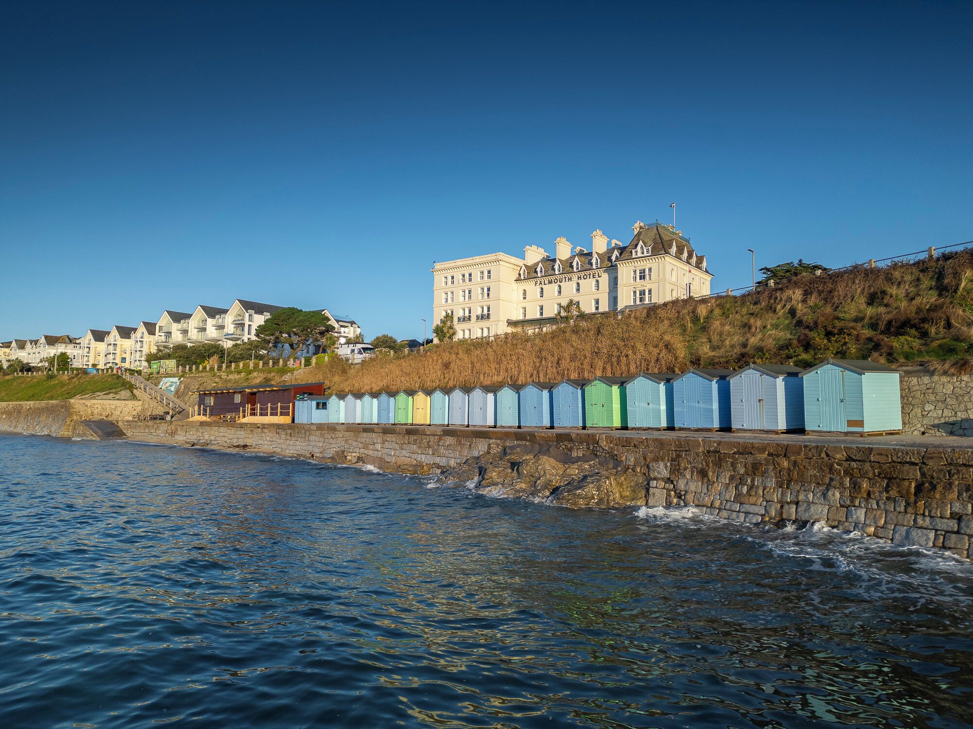 A coastal hotel with a stone wall and colorful beach huts, overlooking the sea.