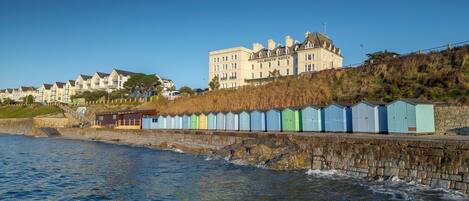 A coastal hotel with a stone wall and colorful beach huts, overlooking the sea.