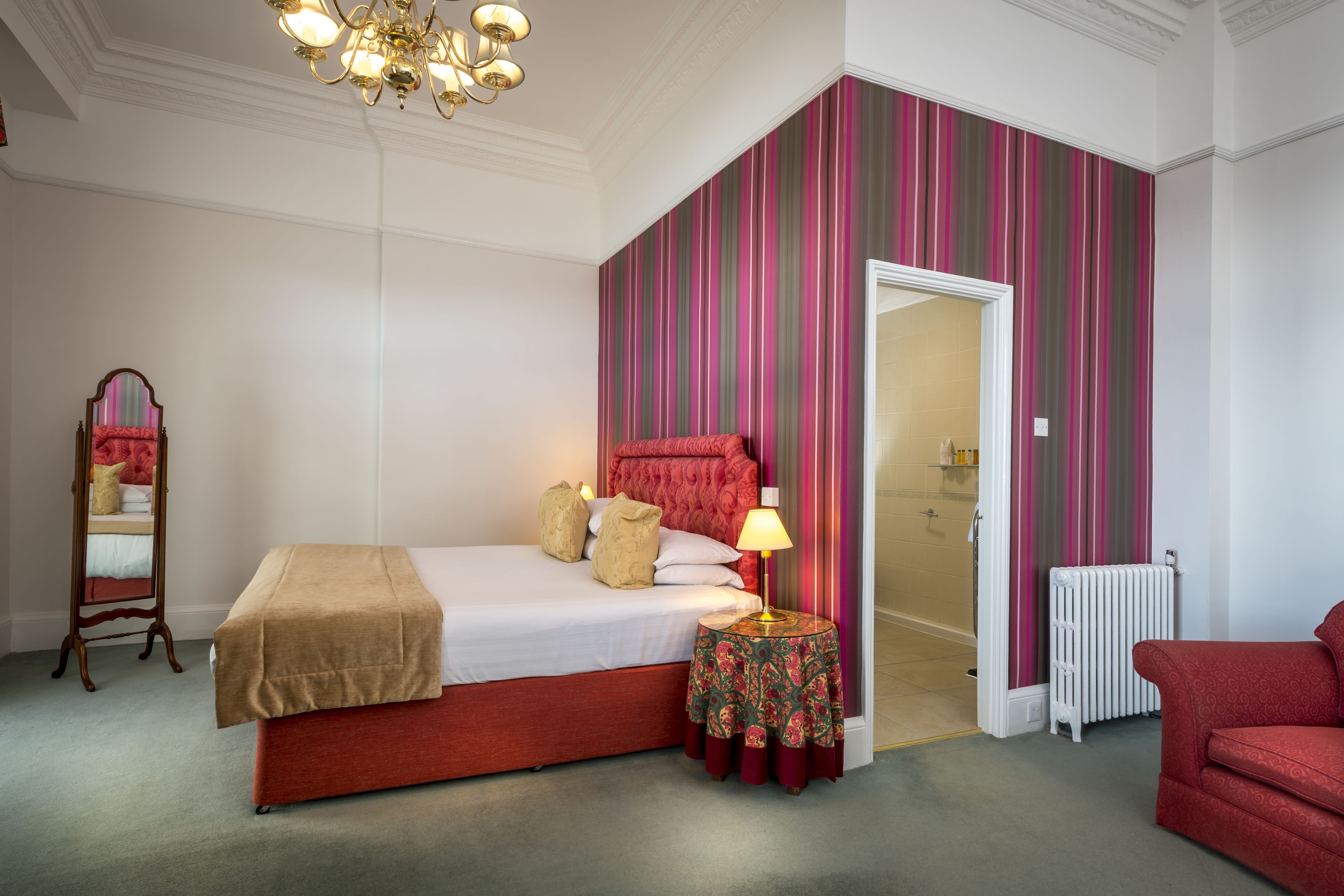 A bedroom with a bed, a bedside table with a lamp, a mirror, a radiator, and a red chair.