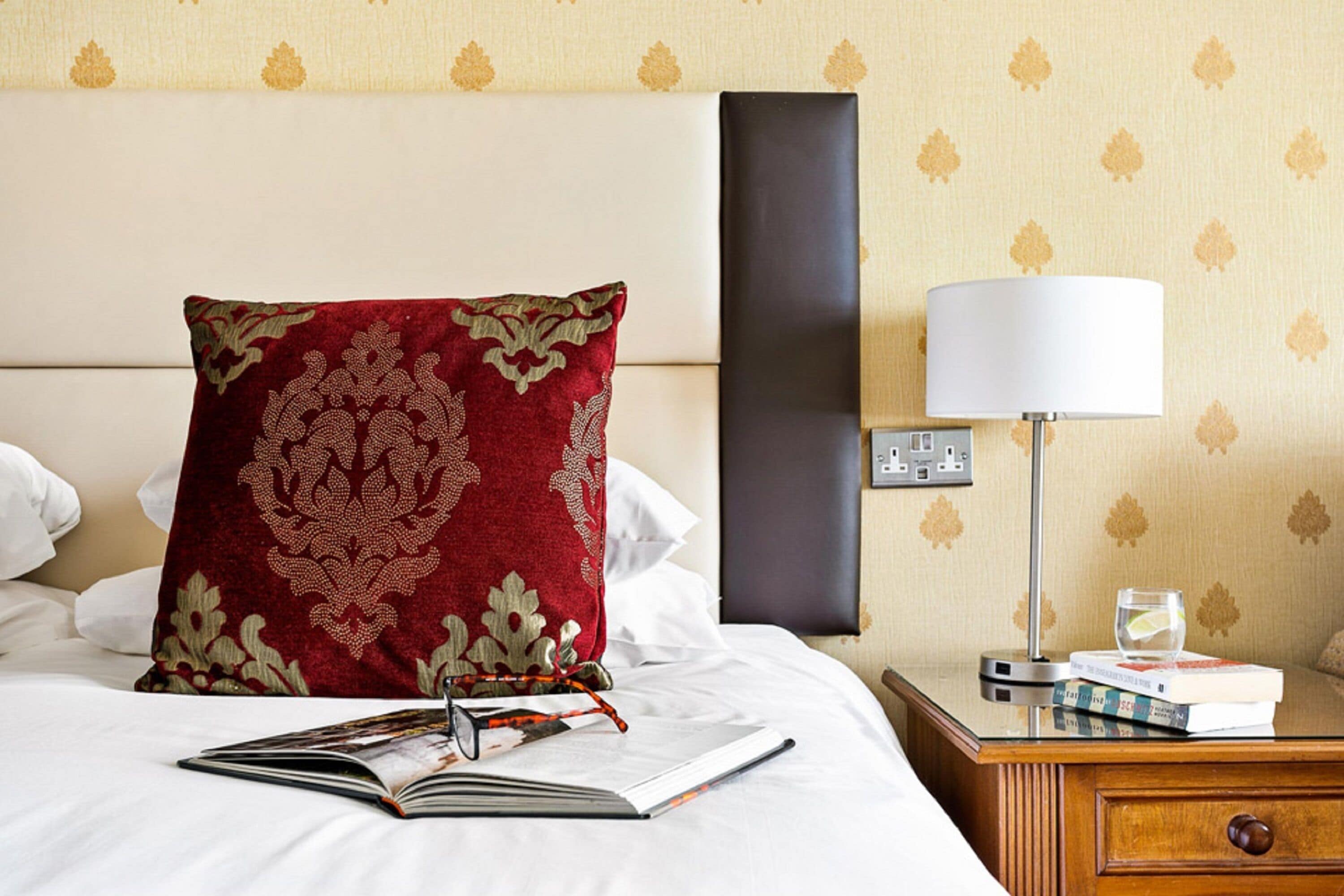 Una cama bien hecha con un cojín estampado en rojo y dorado, un libro y un vaso de agua sobre una mesita de noche.