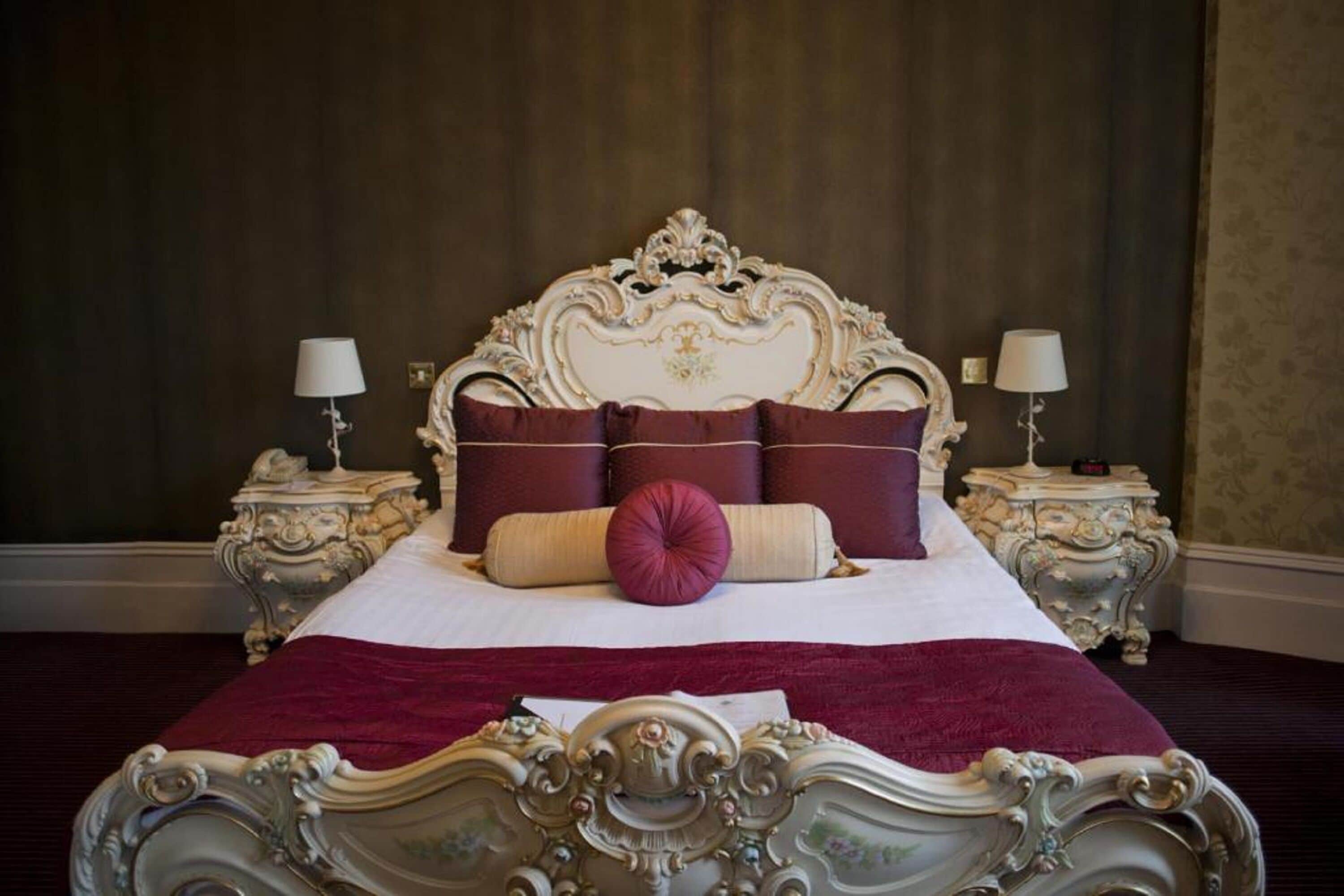 Een klassiek hemelbed met een sierlijk hoofdeinde en voeteneinde, omringd door twee nachtkastjes met lampen, geplaatst tegen een donkere wand.