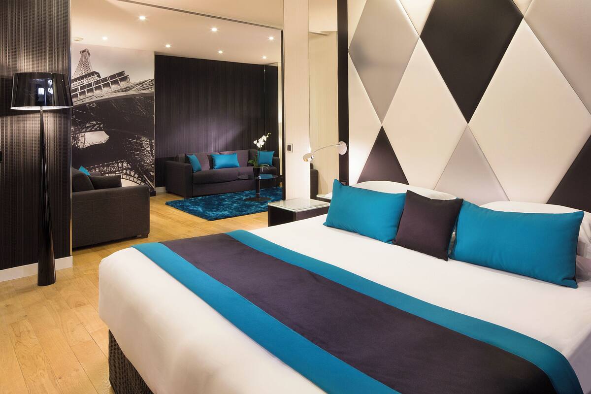 A modern hotel room with a large bed, a sofa, and a small table. There is a wall with a geometric pattern and a mural of a bridge.