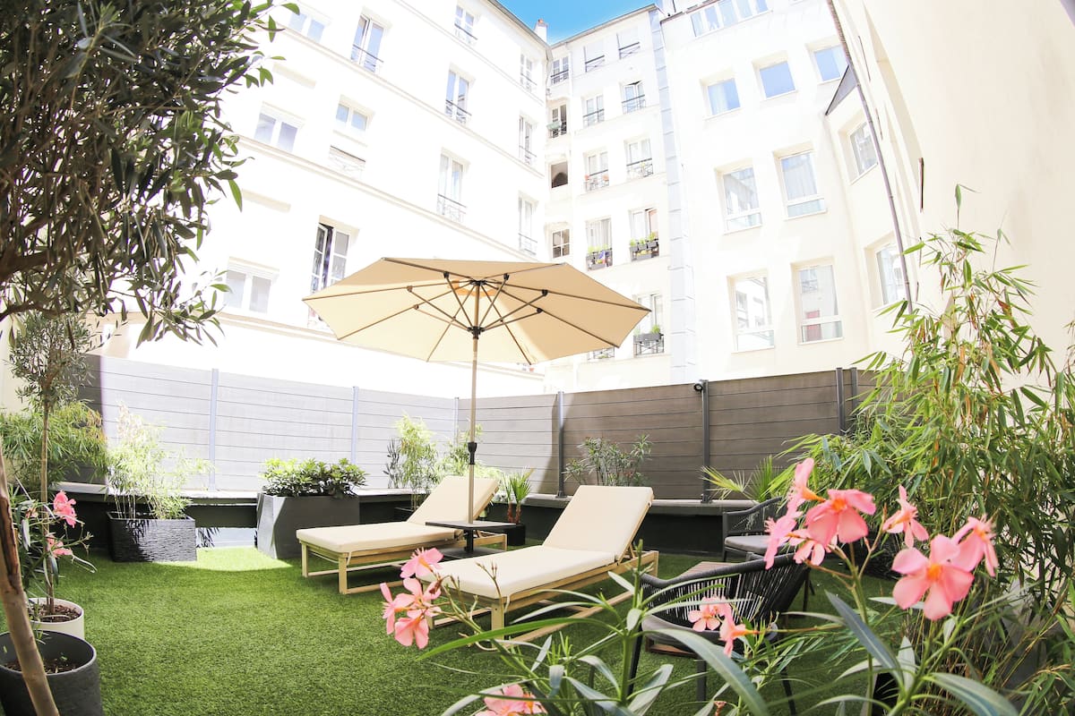 A courtyard with artificial grass, two lounge chairs, a large umbrella, and potted plants.