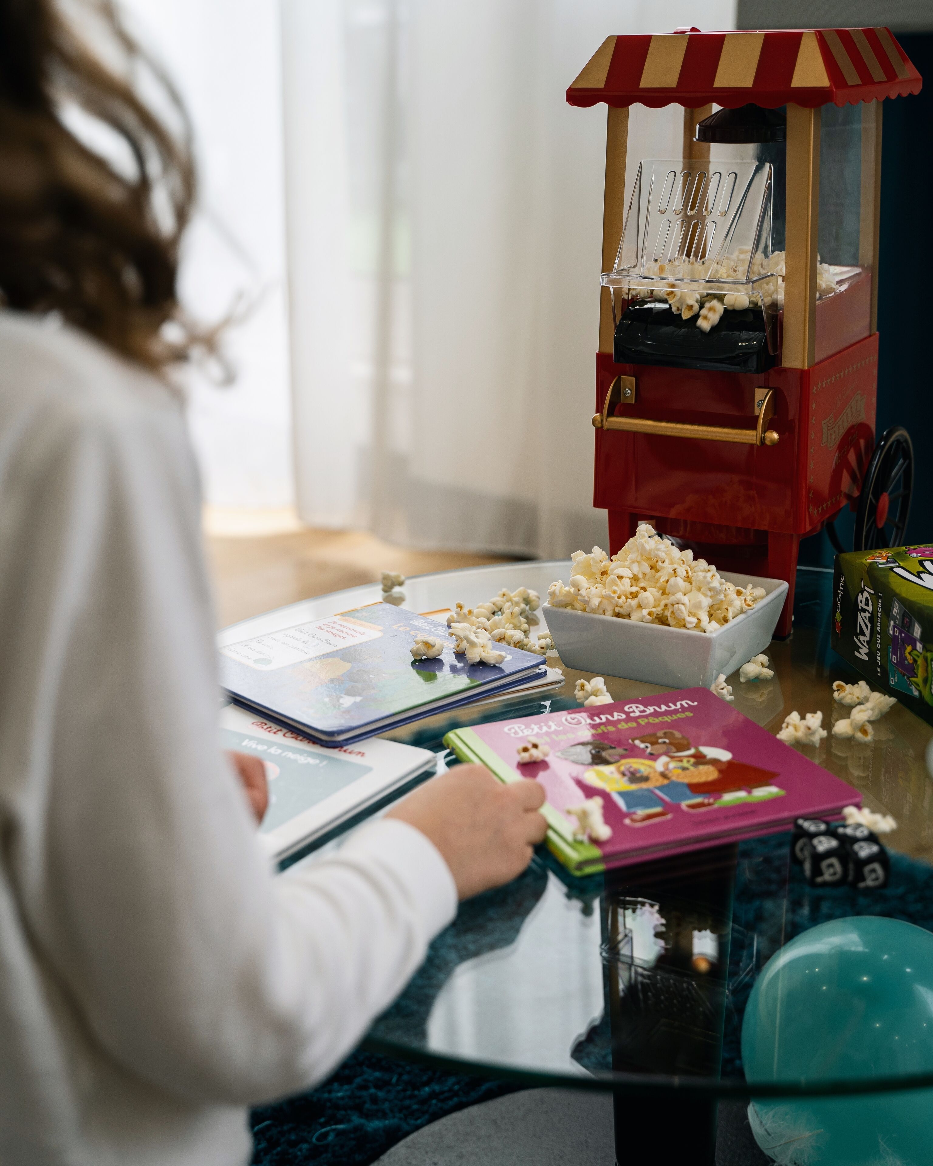 A child sitting at a table with a popcorn machine, books, and dice.