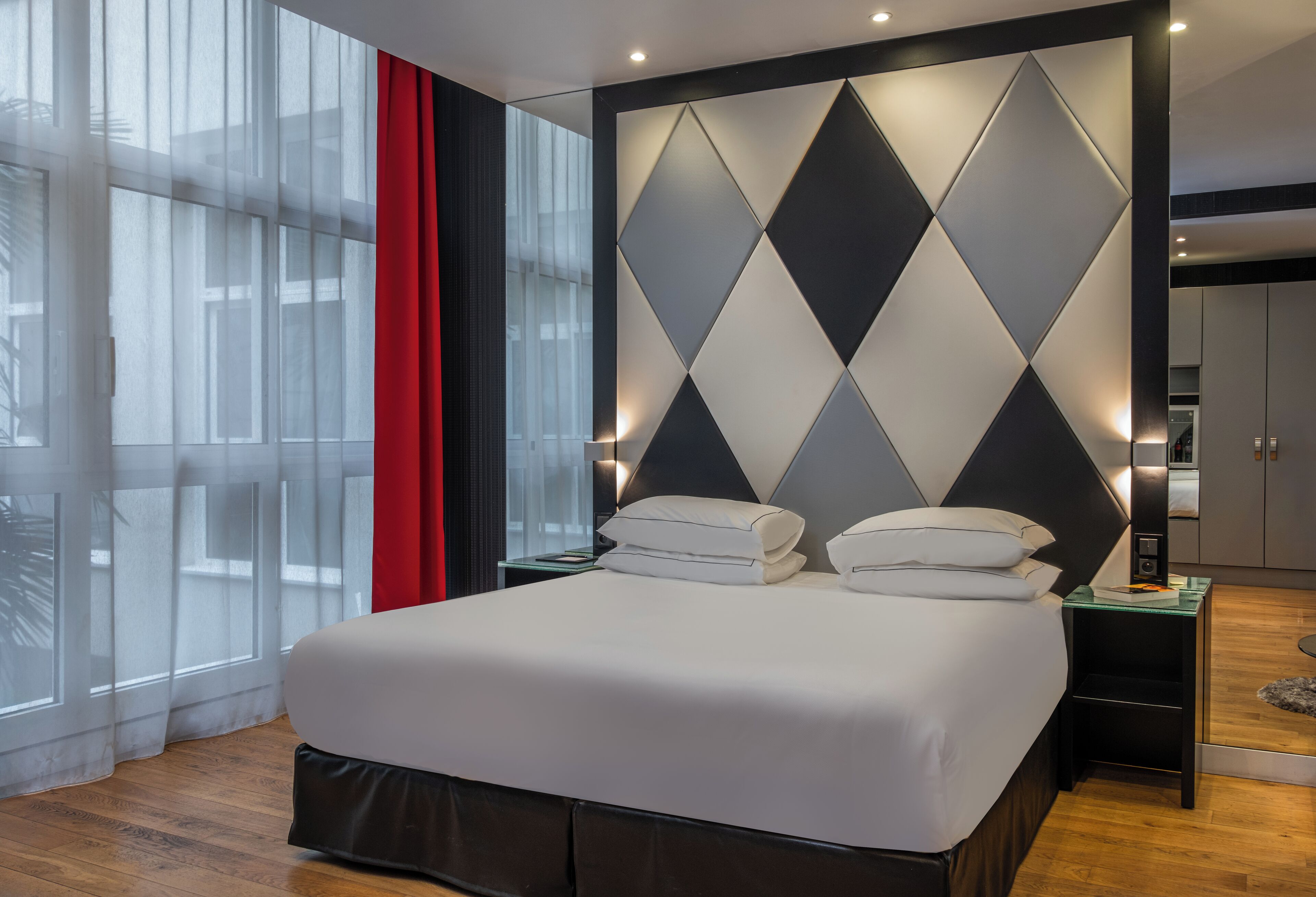 A modern hotel room with a large bed, a geometric wall design, and a view through sheer curtains.