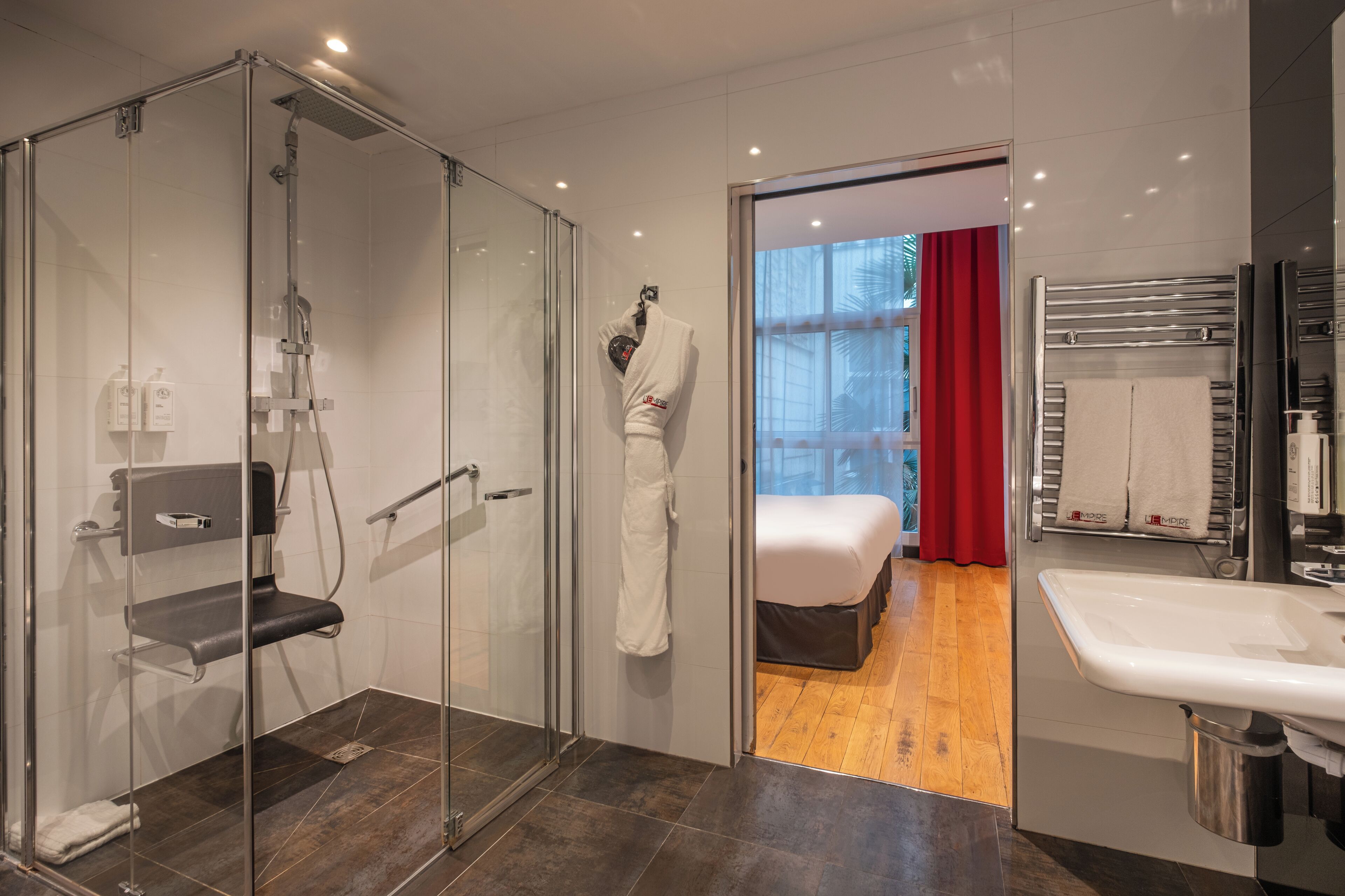 Casa de banho de hotel moderna, com chuveiro em cabine de vidro, pia e cama visível através de uma porta aberta.
