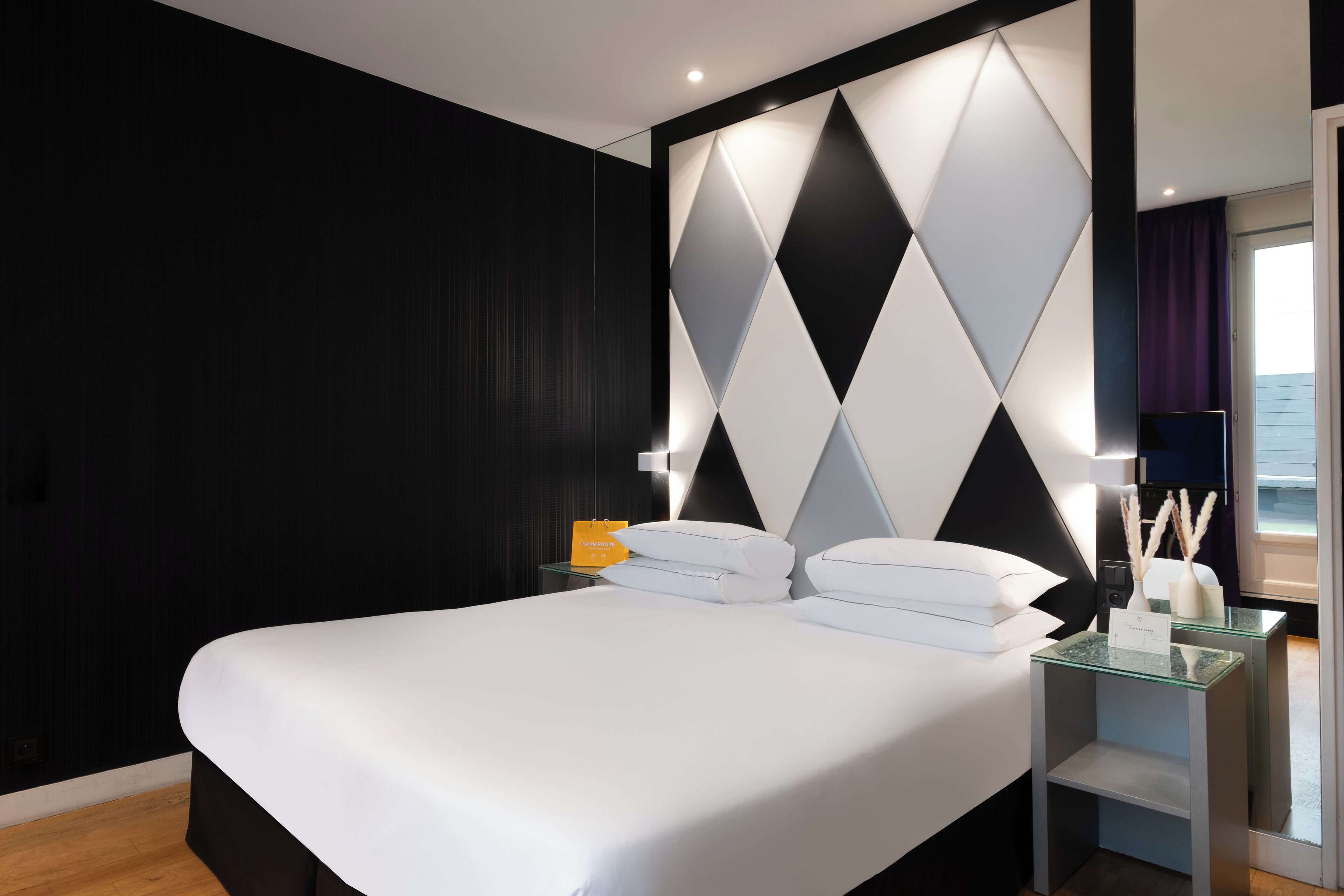 Une chambre d’hôtel moderne avec un grand lit, une table de chevet en verre et un mur orné de motifs géométriques.
