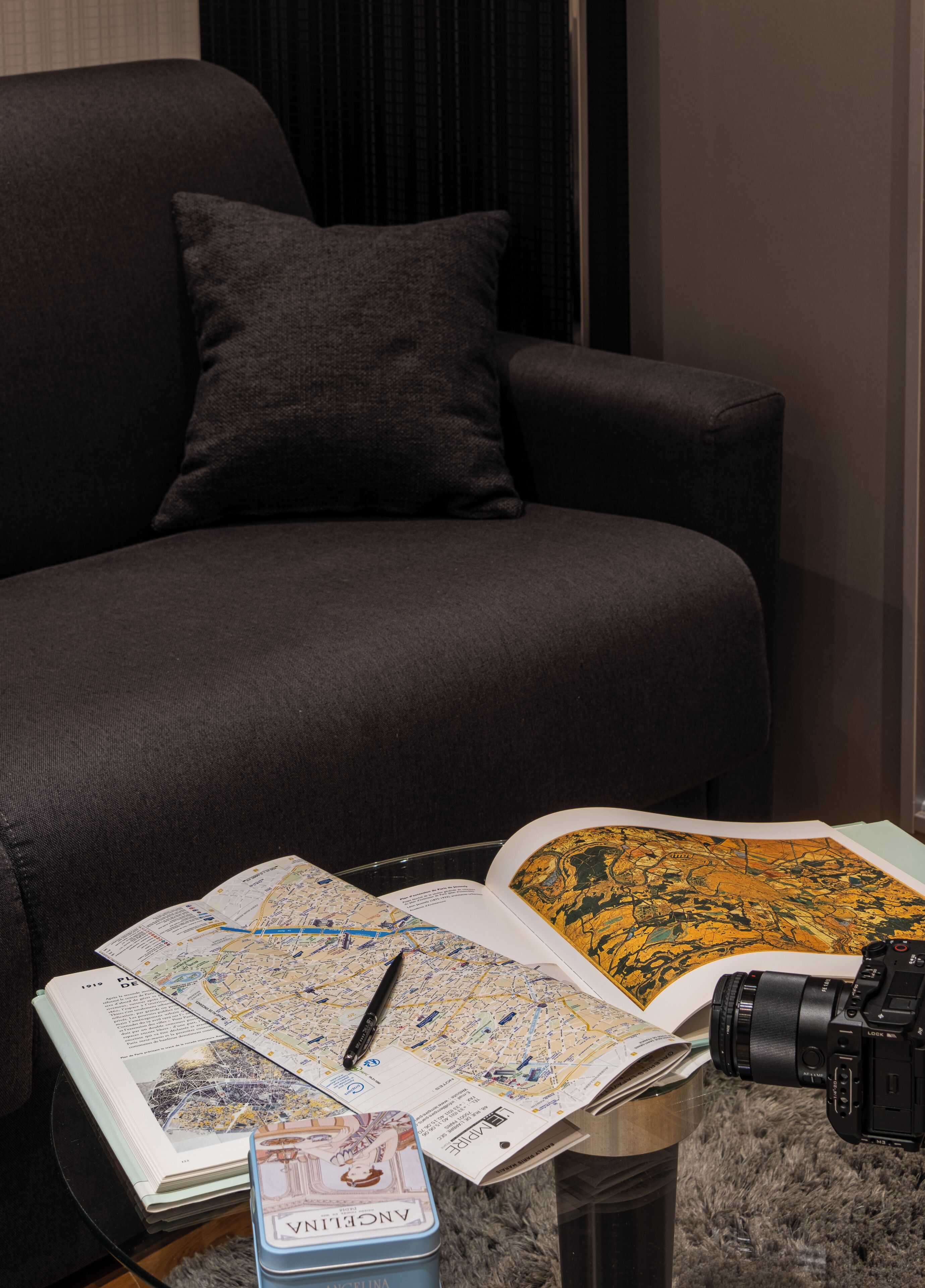 A modern living room with a dark sofa, a coffee table holding a map, a camera, and a tin labeled "ANCIENT."