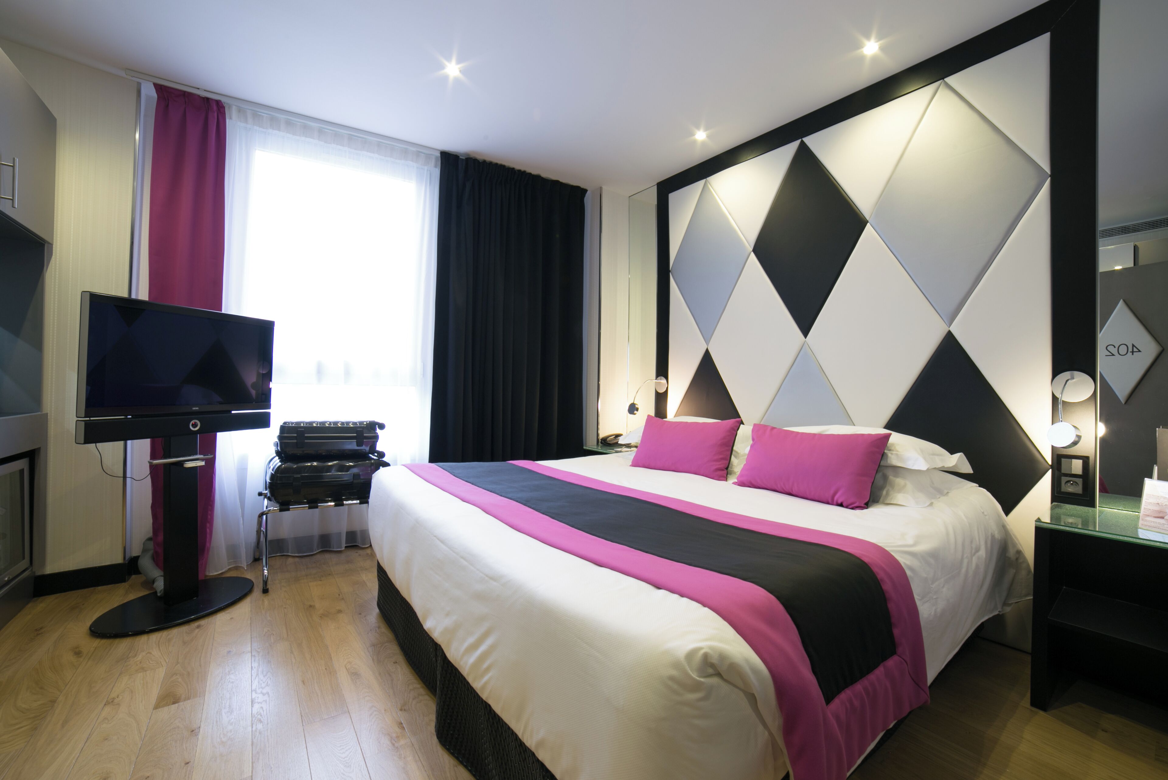 A modern hotel room with a large bed, a flat-screen TV, a desk with a lamp, and a view of the city through the window.