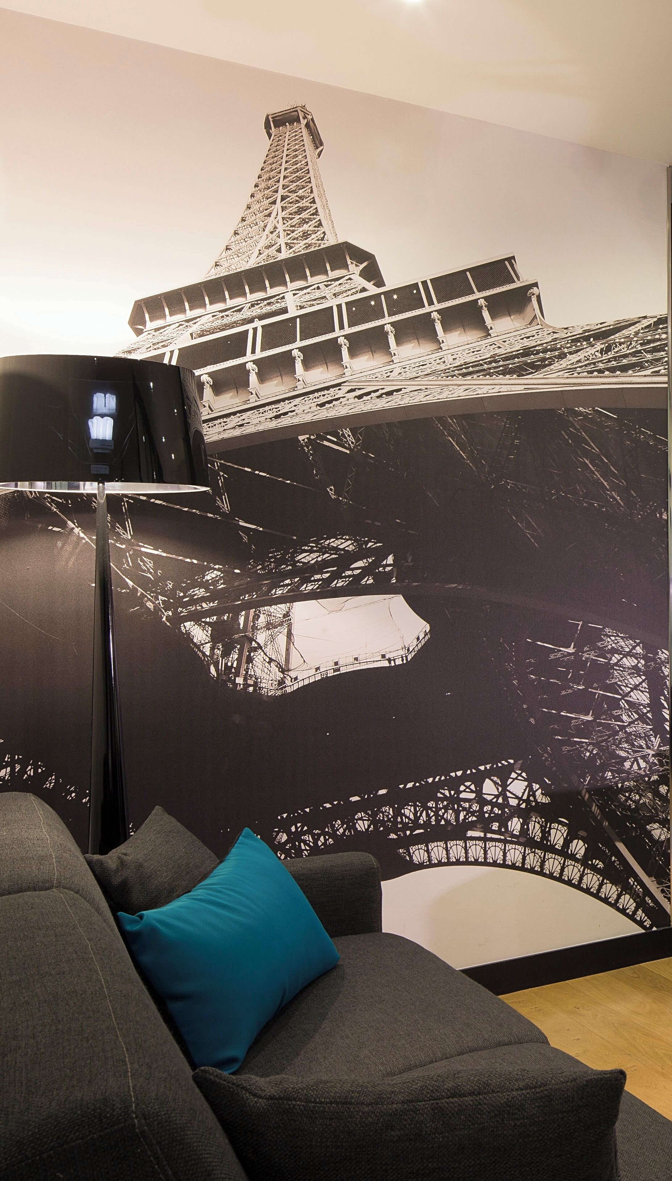 Una habitación con un gran mural de la Torre Eiffel, un sofá negro moderno y una mesita con una lámpara.