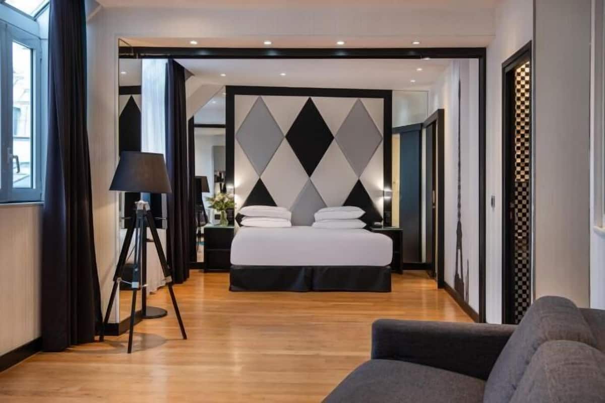 A modern hotel room with a bed, a standing lamp, and a view through an open door.