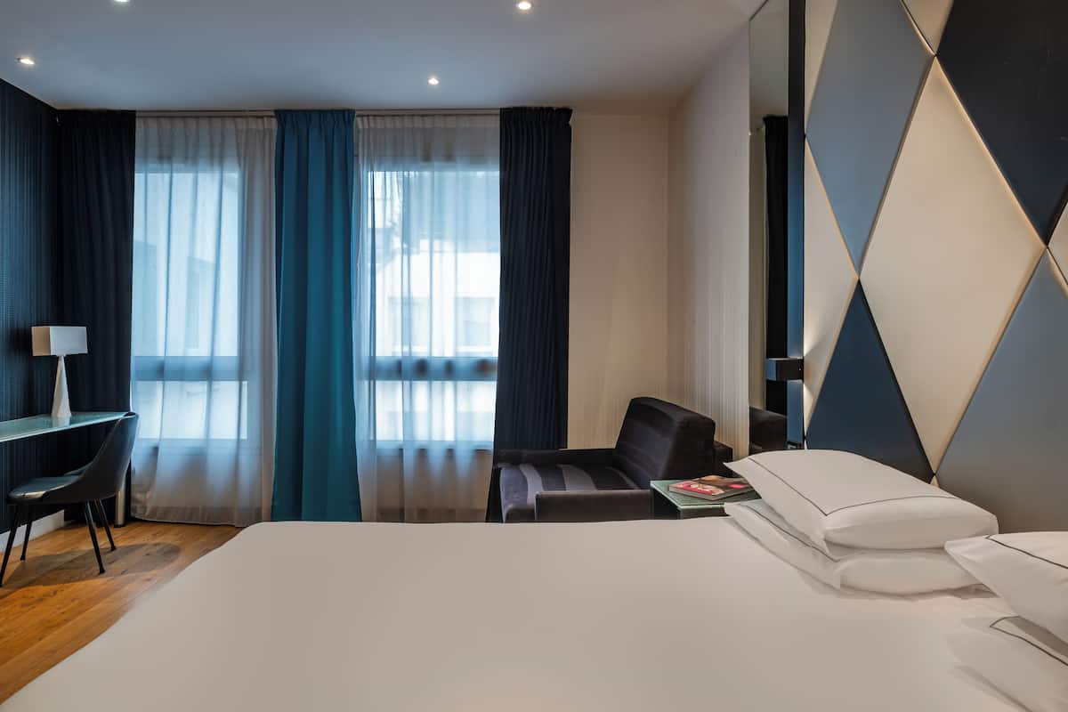 A modern hotel room with a large bed, a desk with a chair, a sofa, and a view of the city through large windows with curtains.