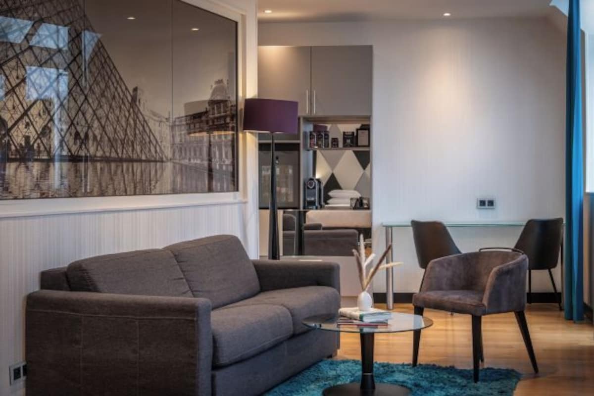 A modern living room with a sofa, armchair, and coffee table. There is a large window with a view of buildings, and a kitchen area is visible in the background.