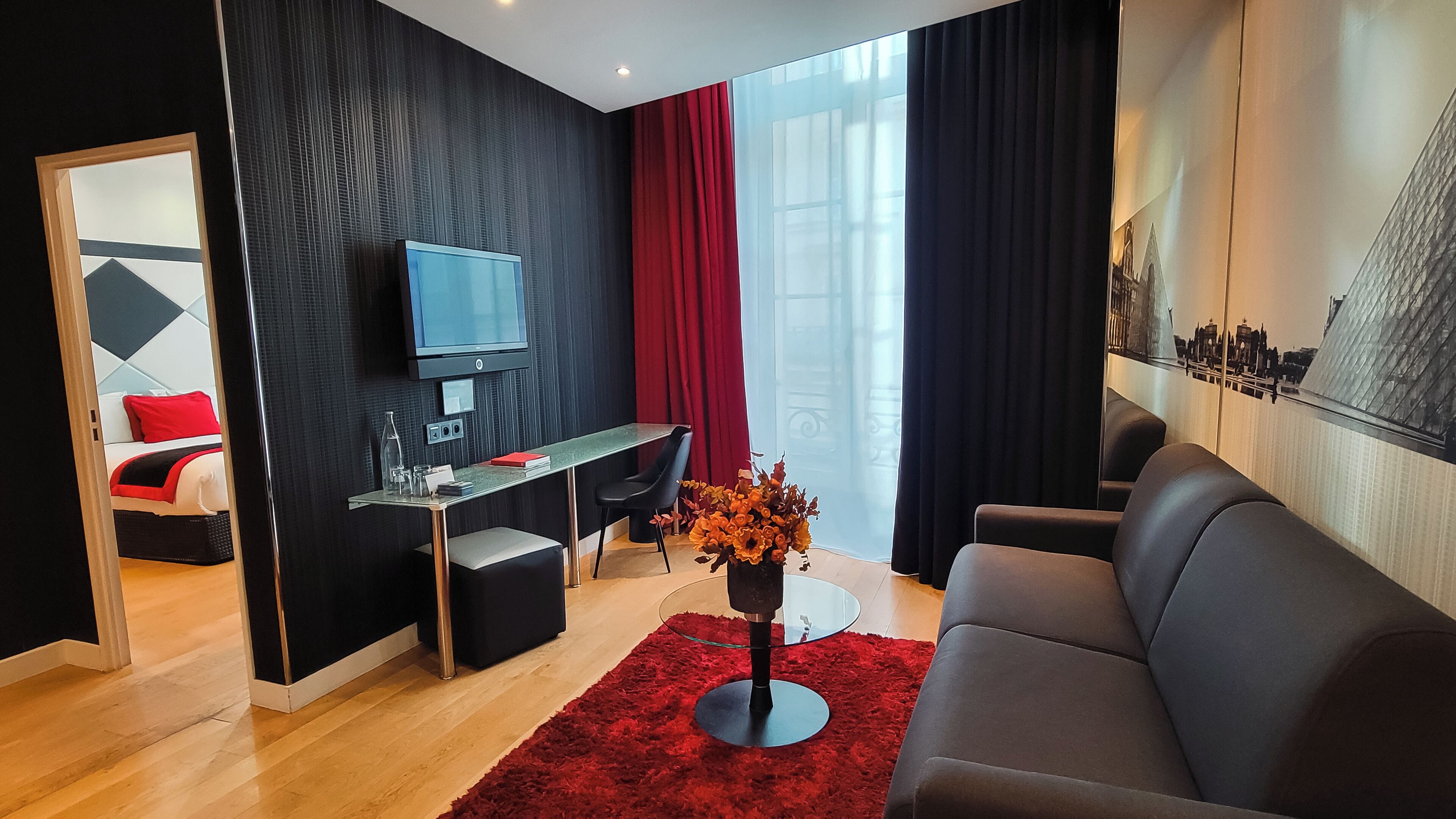 A modern living room with a black sofa, a glass coffee table, a TV, a desk with a chair, and a red rug.